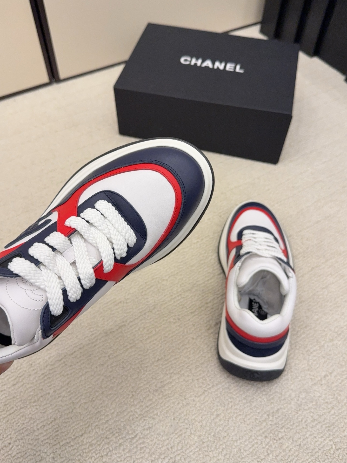A pair of Chanel tricolor sneakers in white, navy blue, and red with a chunky sole and the CC logo, displayed next to a black Chanel shoe box. - Hình 6