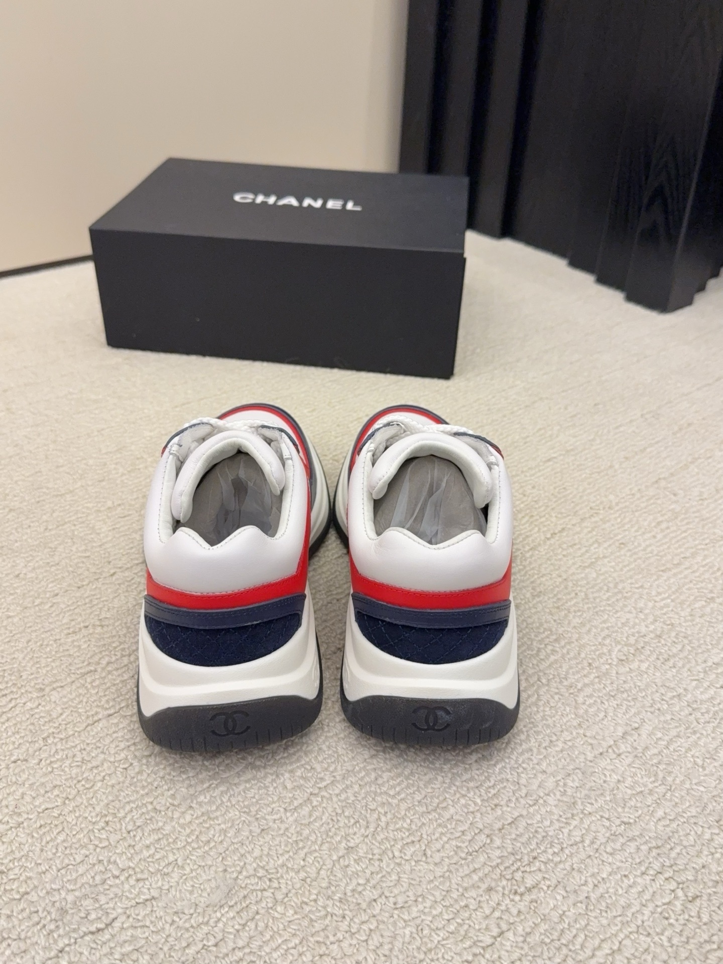 A pair of Chanel tricolor sneakers in white, navy blue, and red with a chunky sole and the CC logo, displayed next to a black Chanel shoe box. - Hình 4