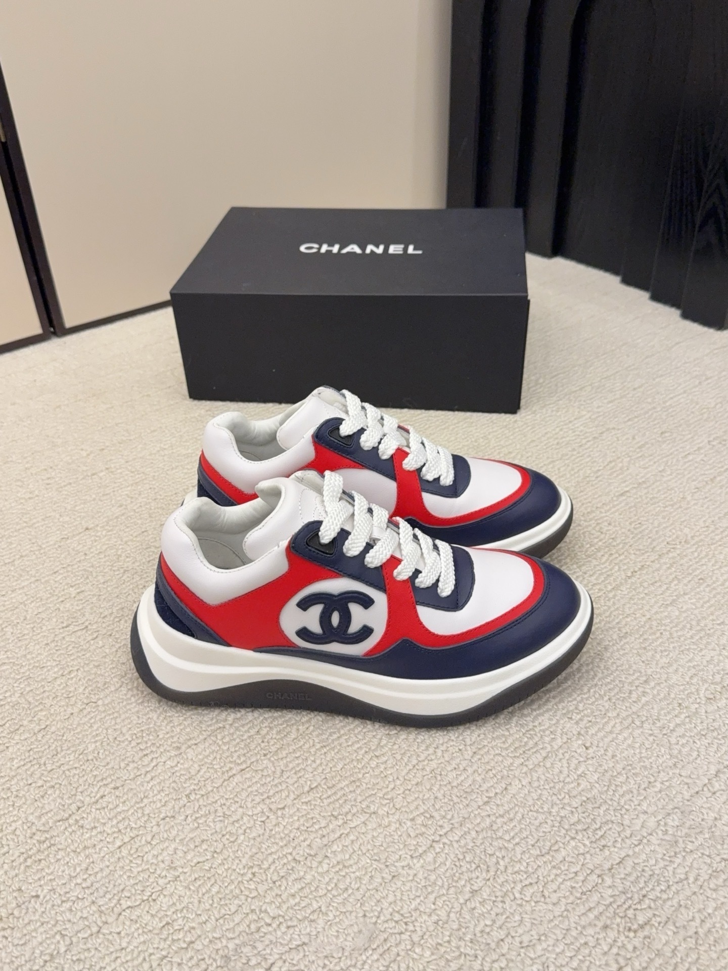 A pair of Chanel tricolor sneakers in white, navy blue, and red with a chunky sole and the CC logo, displayed next to a black Chanel shoe box. - Hình 3