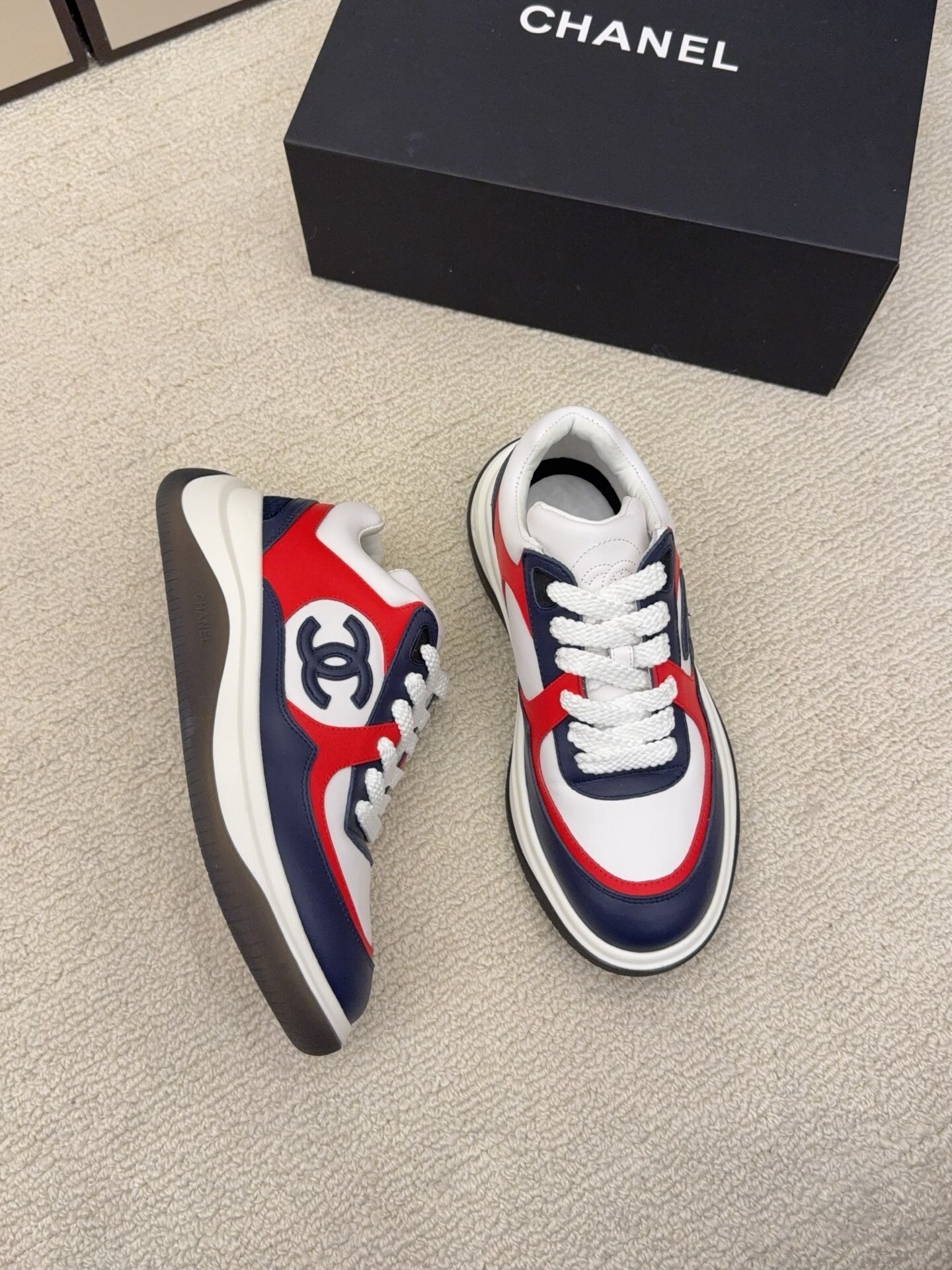 A pair of Chanel tricolor sneakers in white, navy blue, and red with a chunky sole and the CC logo, displayed next to a black Chanel shoe box. - Hình 2