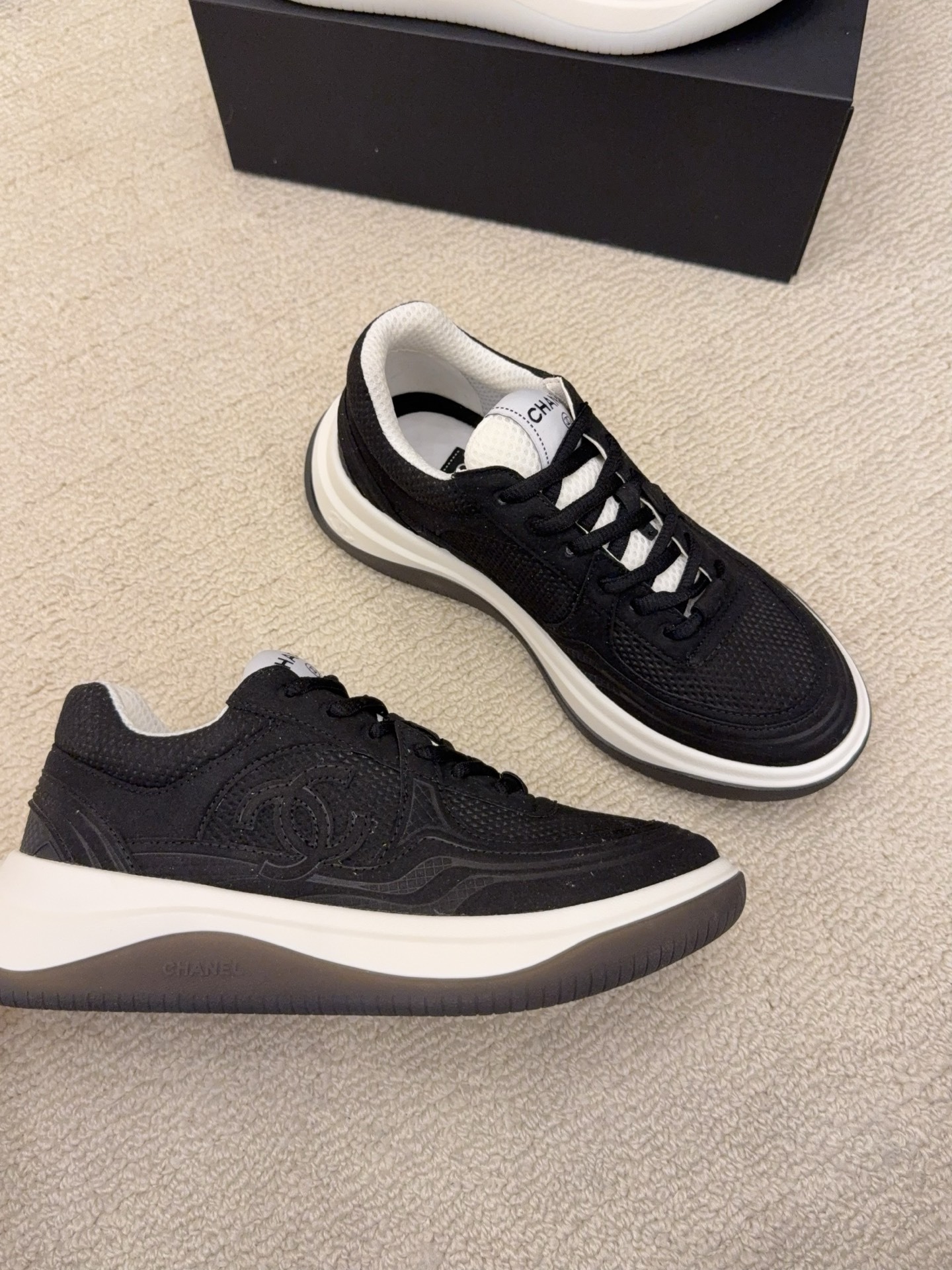 A pair of black and a pair of beige Chanel luxury platform sneakers with mesh detailing and interlocking CC logo displayed on a carpeted floor. - Hình 3