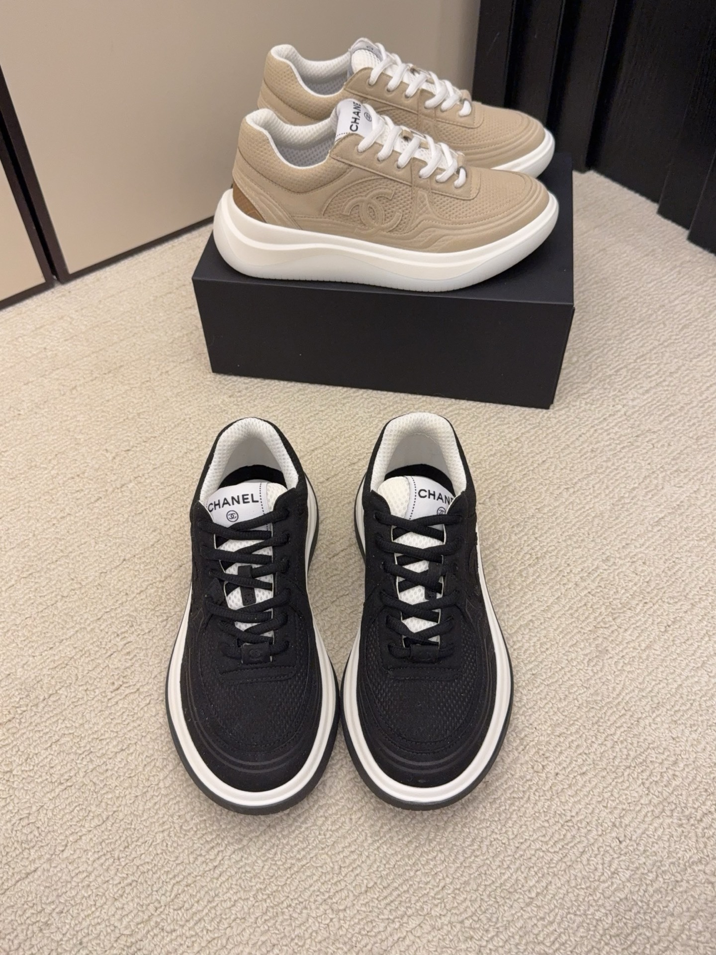 A pair of black and a pair of beige Chanel luxury platform sneakers with mesh detailing and interlocking CC logo displayed on a carpeted floor. - Hình 2