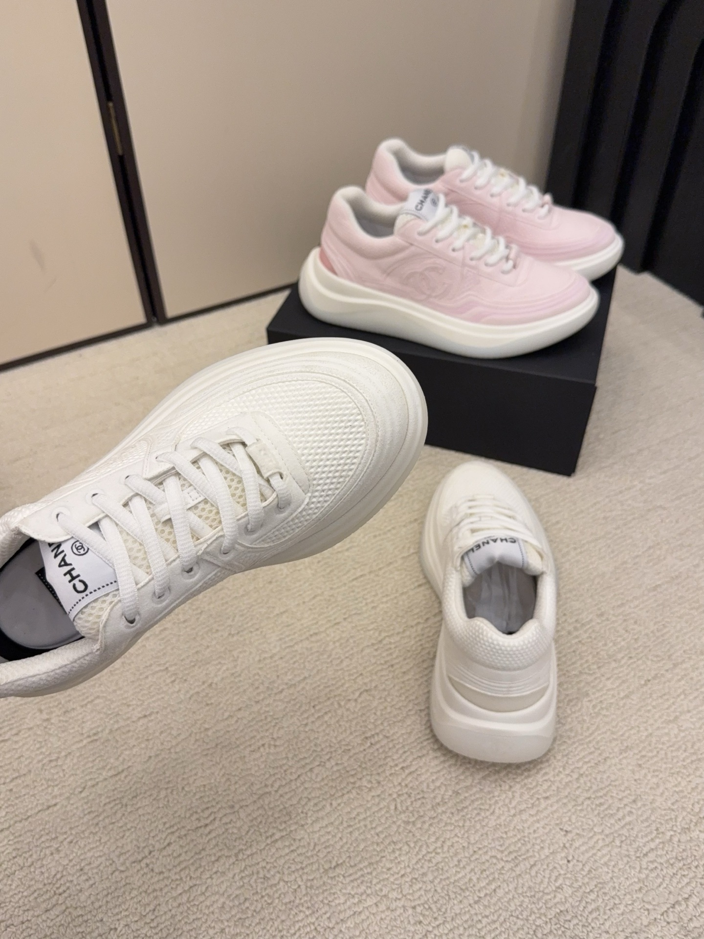 Two pairs of Chanel luxury platform sneakers in white and pastel pink colors displayed on a neutral carpet next to a black shoe box. - Hình 8