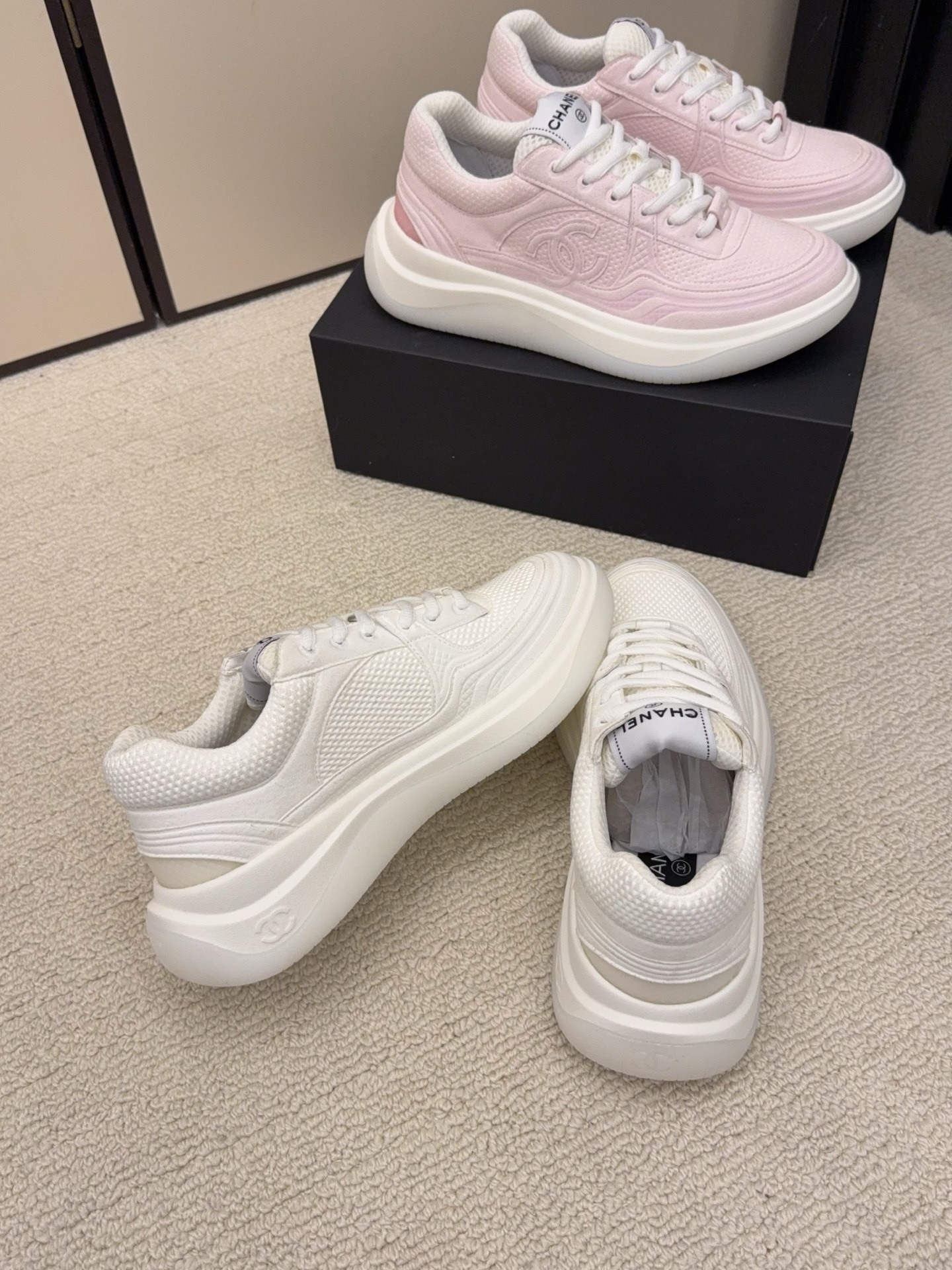 Two pairs of Chanel luxury platform sneakers in white and pastel pink colors displayed on a neutral carpet next to a black shoe box. - Hình 7