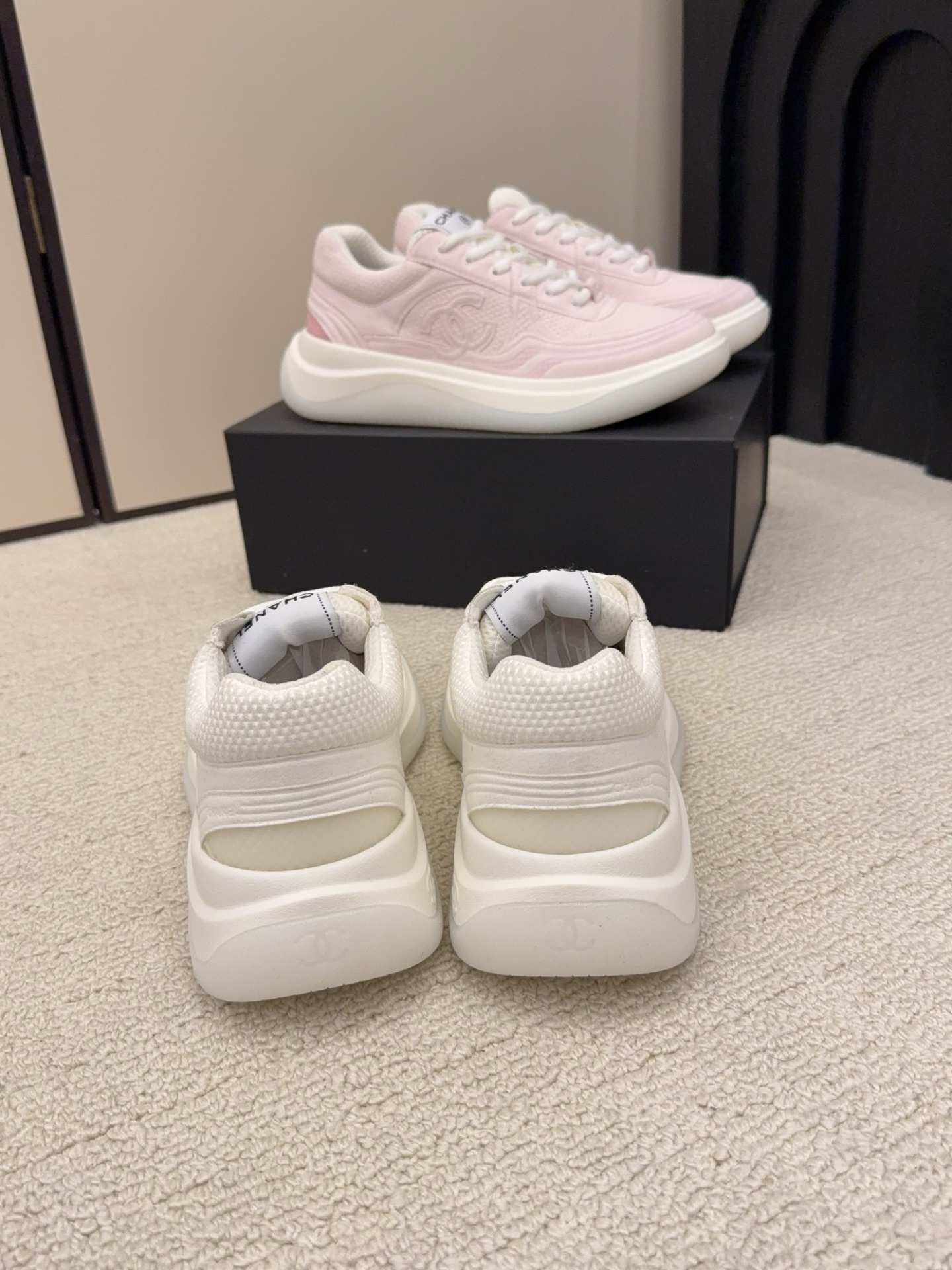 Two pairs of Chanel luxury platform sneakers in white and pastel pink colors displayed on a neutral carpet next to a black shoe box. - Hình 6