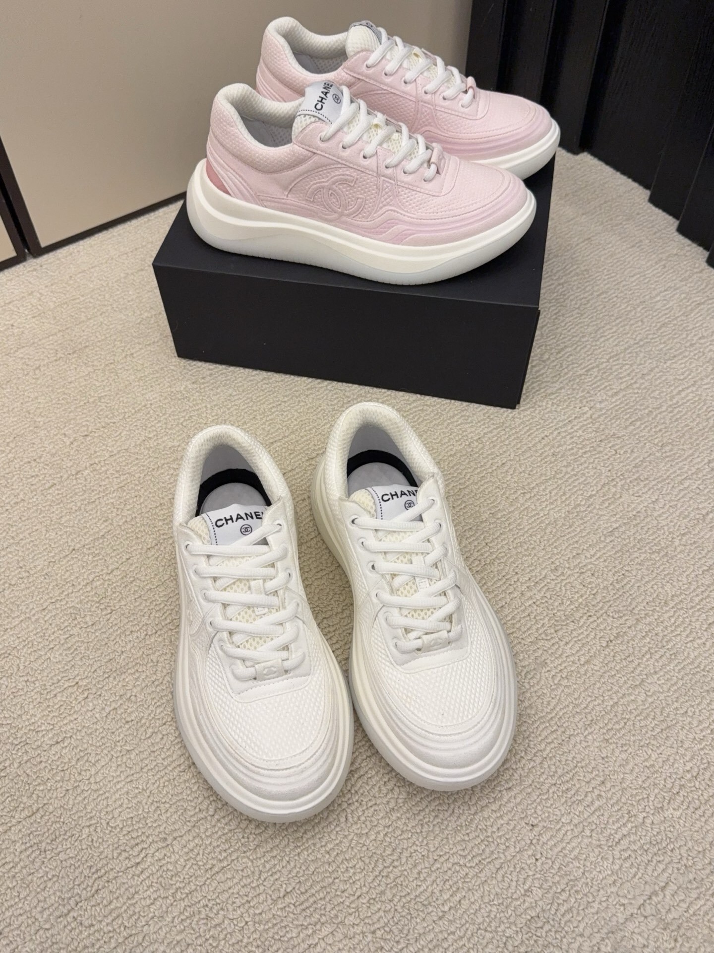 Two pairs of Chanel luxury platform sneakers in white and pastel pink colors displayed on a neutral carpet next to a black shoe box. - Hình 5