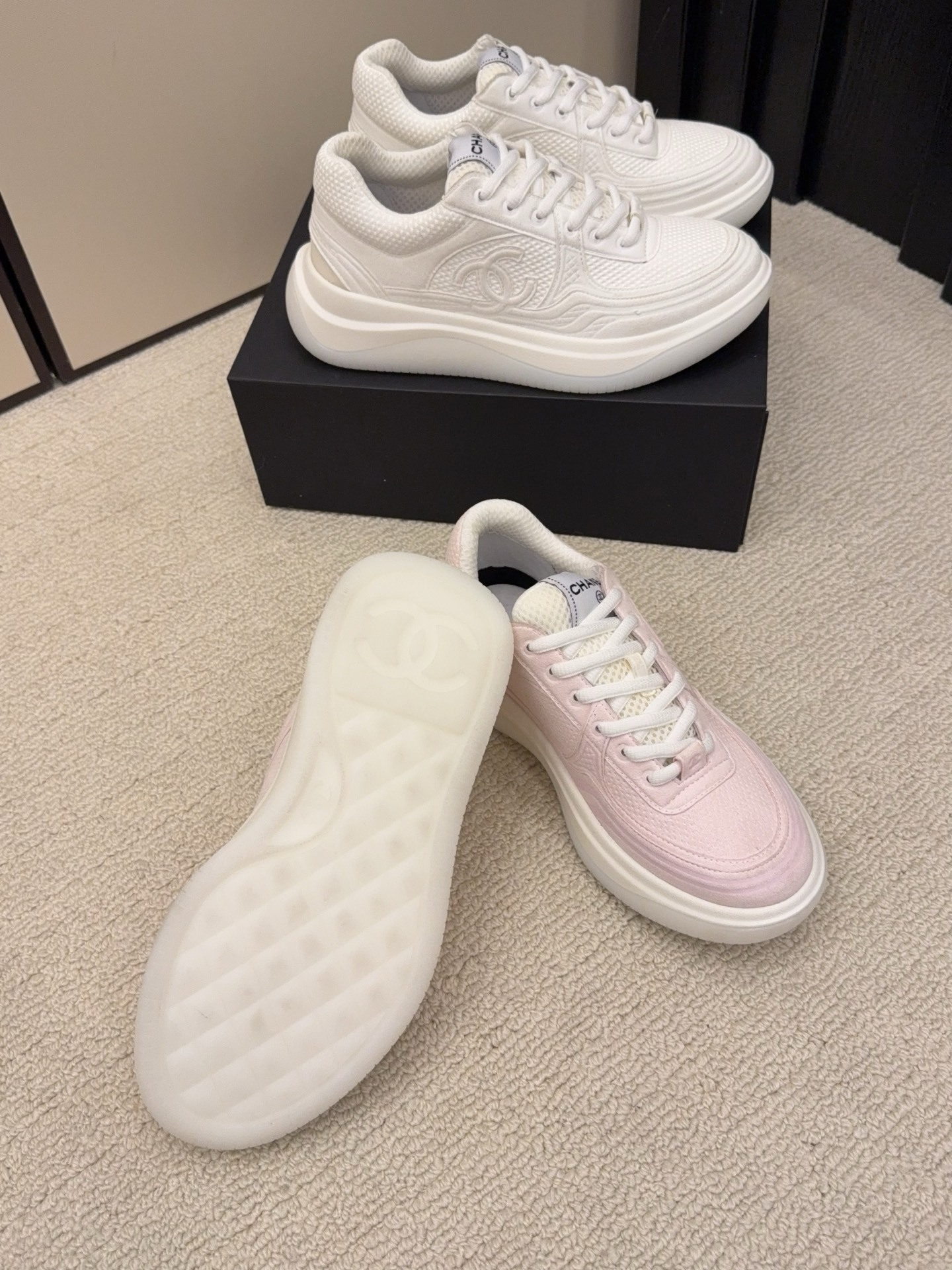Two pairs of Chanel luxury platform sneakers in white and pastel pink colors displayed on a neutral carpet next to a black shoe box. - Hình 4