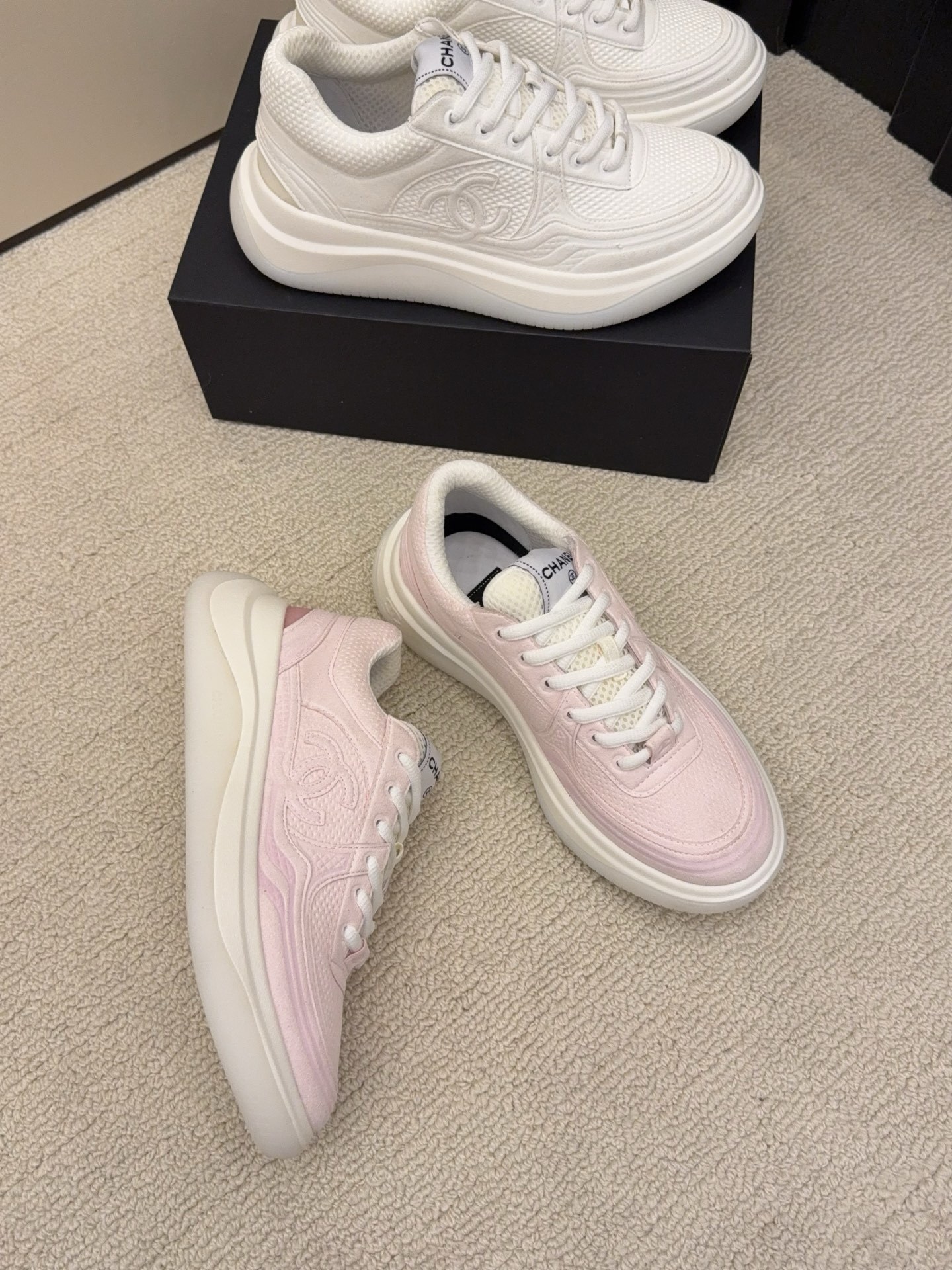 Two pairs of Chanel luxury platform sneakers in white and pastel pink colors displayed on a neutral carpet next to a black shoe box. - Hình 3