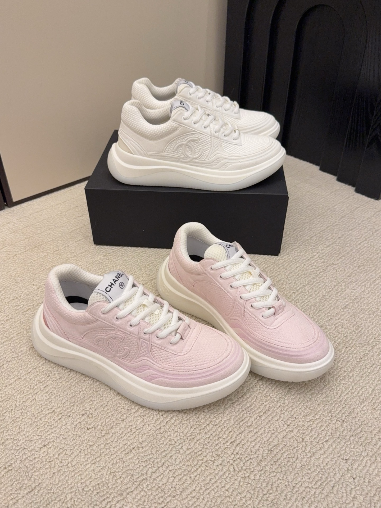 Two pairs of Chanel luxury platform sneakers in white and pastel pink colors displayed on a neutral carpet next to a black shoe box. - Hình 2