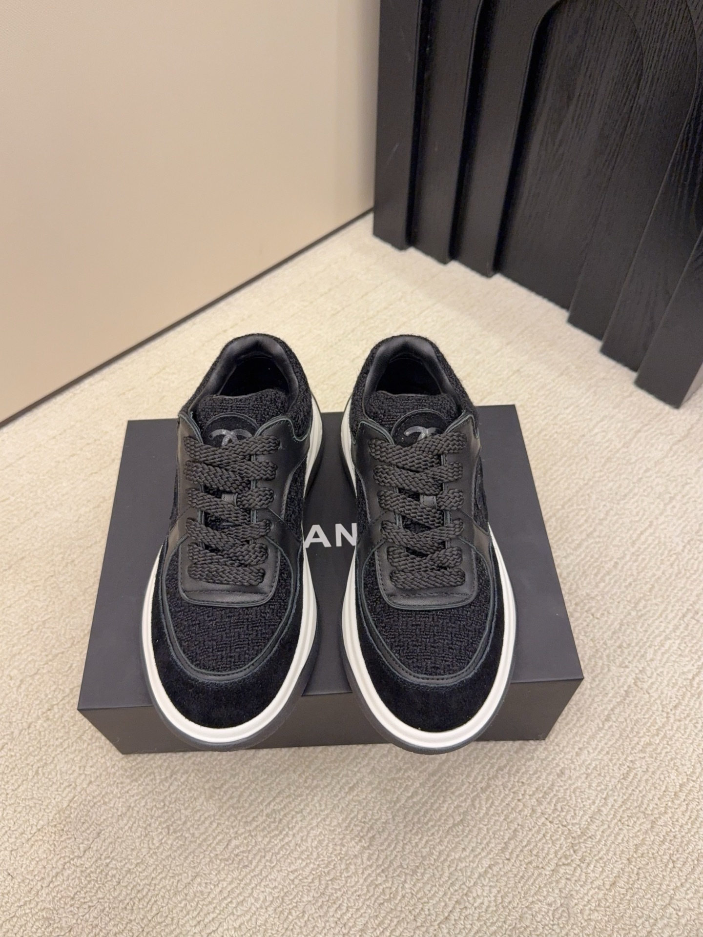 A pair of luxury black Chanel sneakers made of tweed and leather with white chunky soles on a black box. - Hình 2
