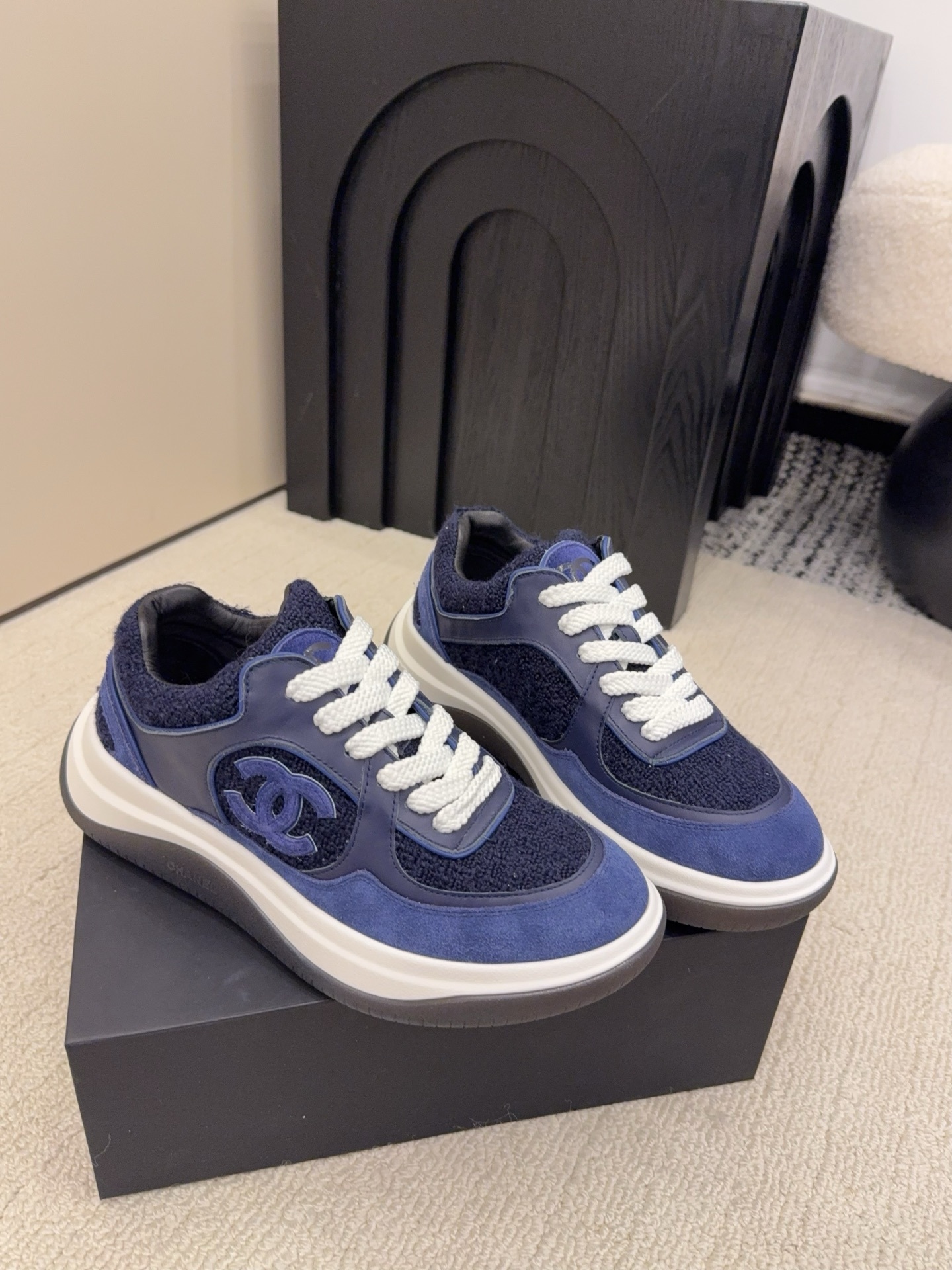 A pair of Chanel navy blue sneakers featuring tweed and suede panels with white laces on a Chanel shoe box. - Hình 4