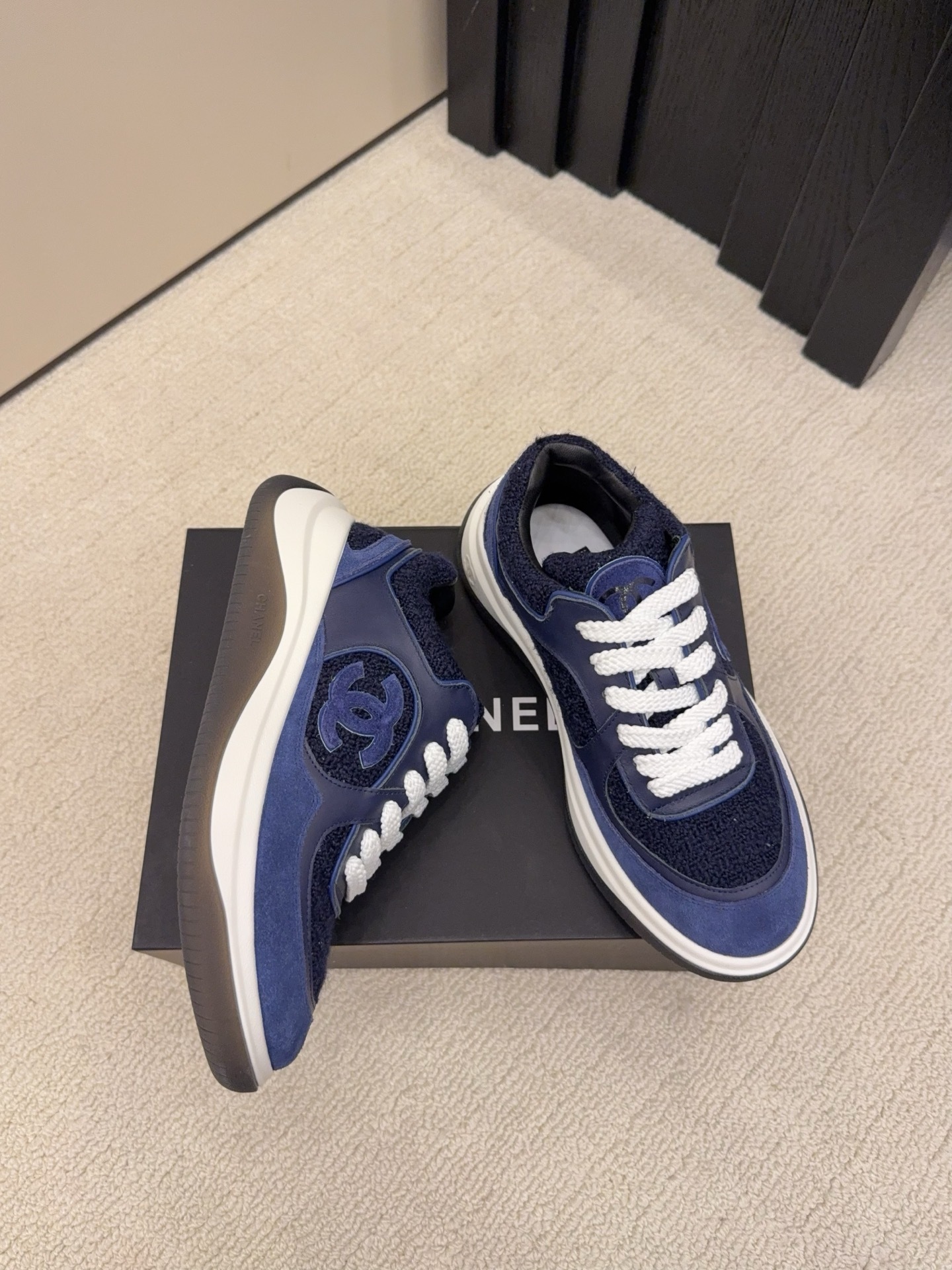 A pair of Chanel navy blue sneakers featuring tweed and suede panels with white laces on a Chanel shoe box. - Hình 3