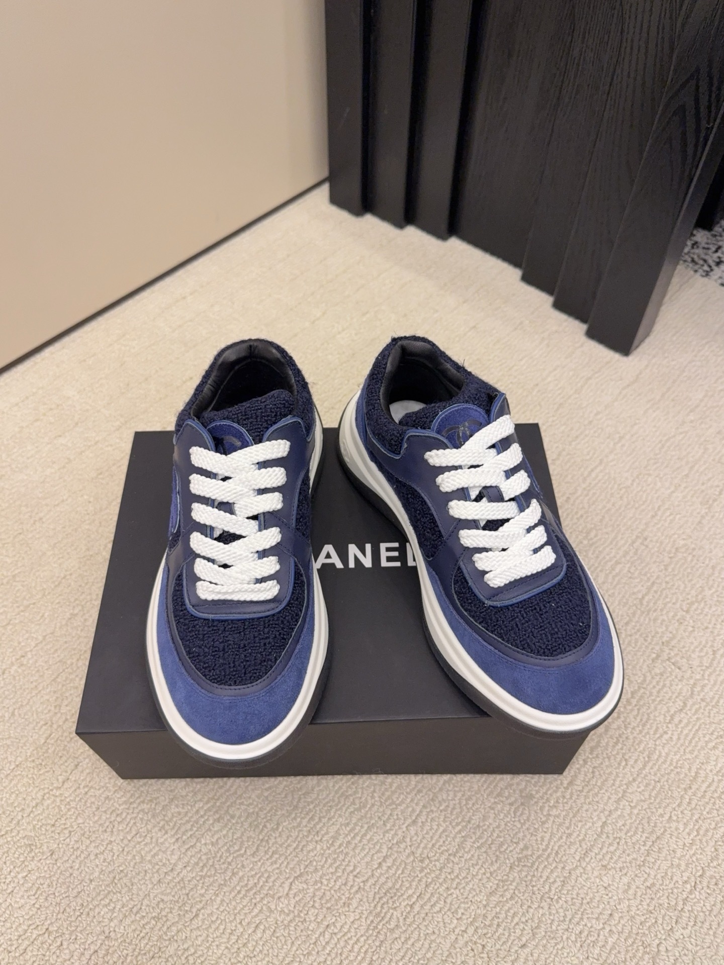 A pair of Chanel navy blue sneakers featuring tweed and suede panels with white laces on a Chanel shoe box. - Hình 2