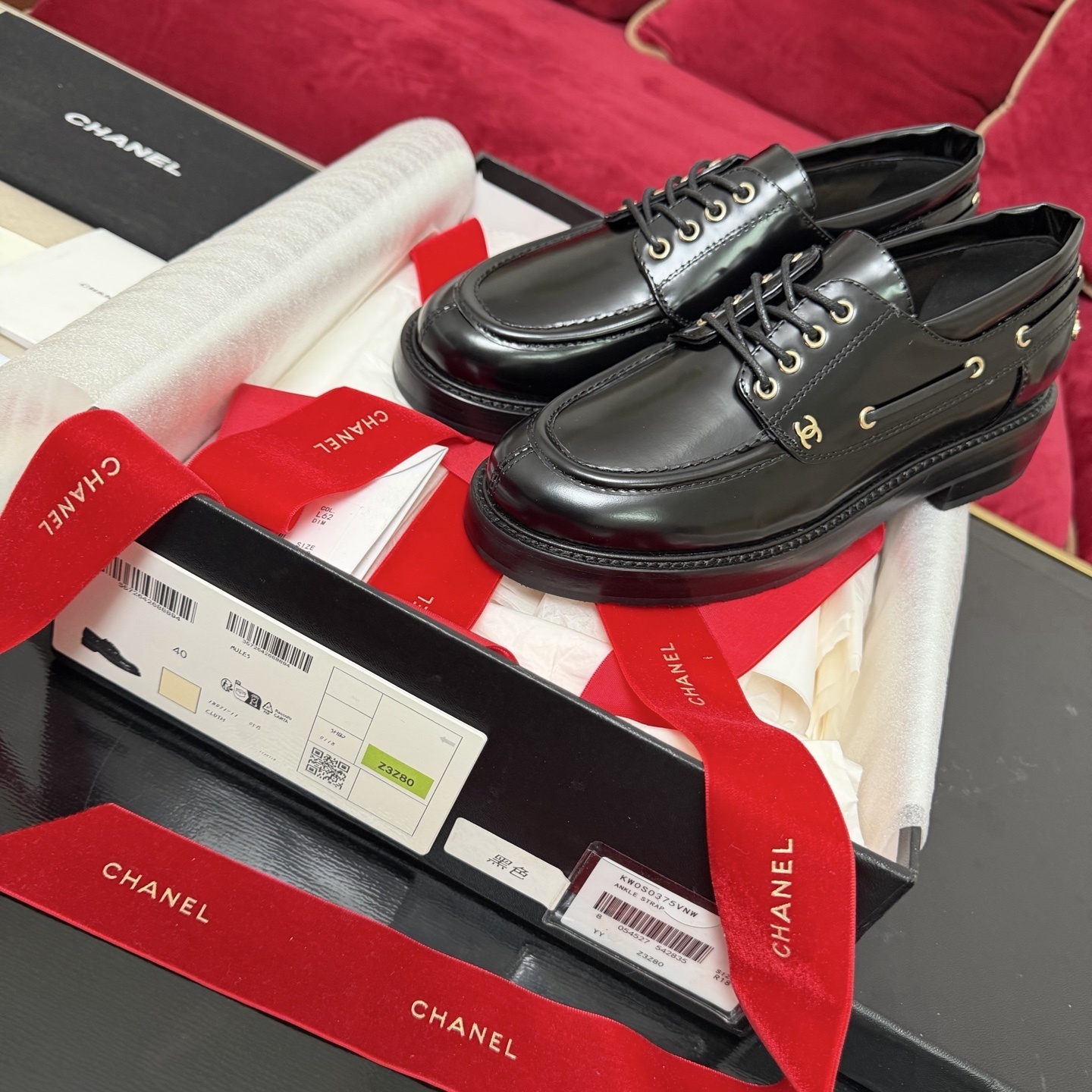 A pair of Chanel black leather lace-up loafers with gold CC logo hardware, displayed in a box with red velvet ribbons. - Hình 2