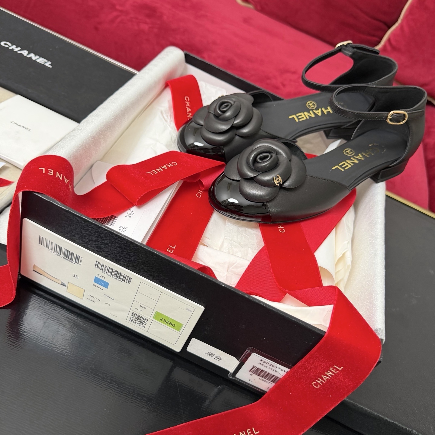 Pair of black Chanel ankle strap flats featuring a large camellia flower and patent leather toe cap in a luxury gift box. - Hình 2