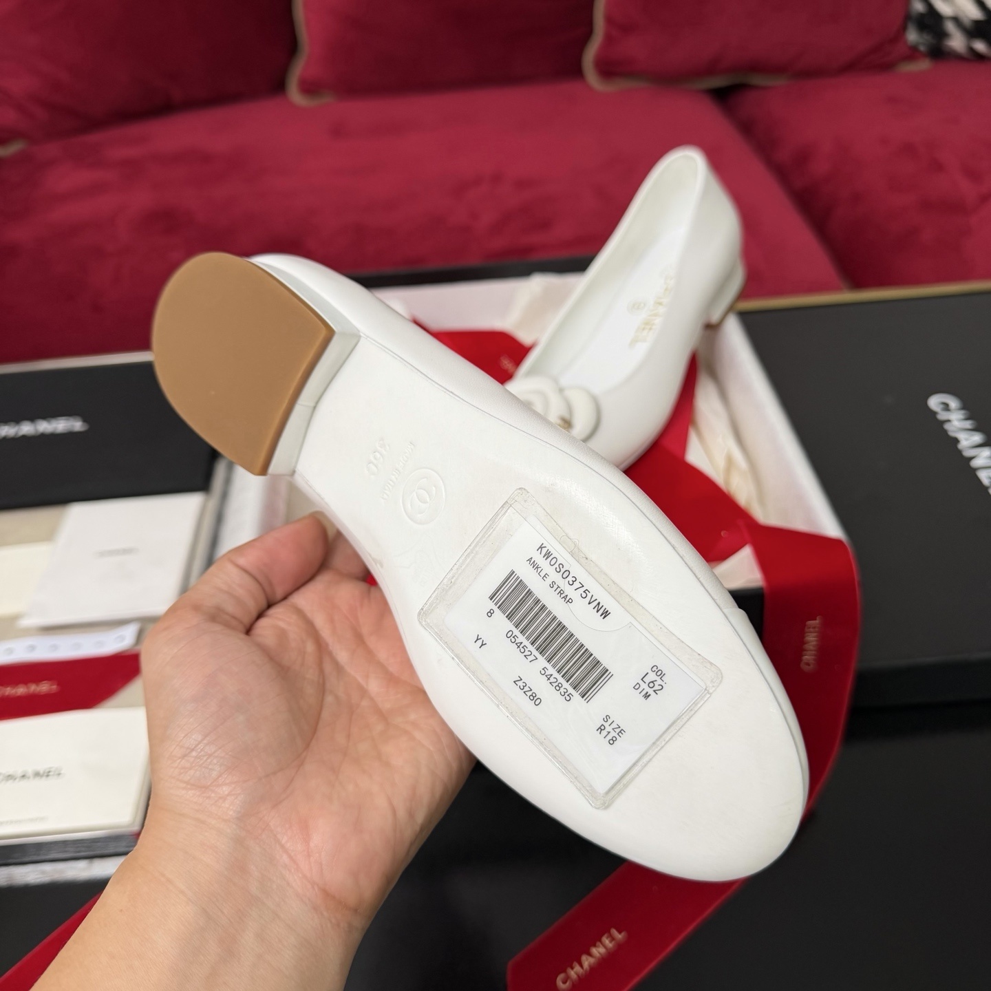 Close-up of Chanel white ballet flats featuring a leather camellia flower and gold CC logo inside a luxury gift box with red ribbon. - Hình 9