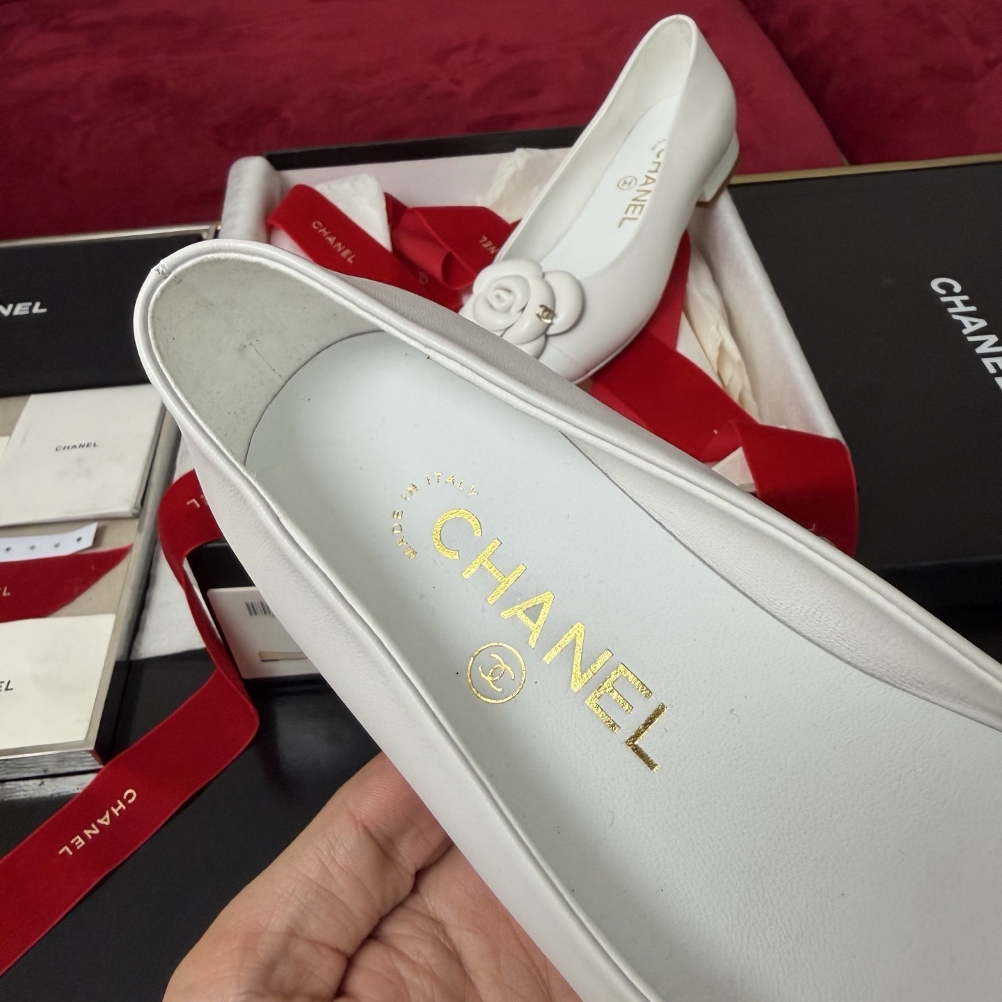 Close-up of Chanel white ballet flats featuring a leather camellia flower and gold CC logo inside a luxury gift box with red ribbon. - Hình 8