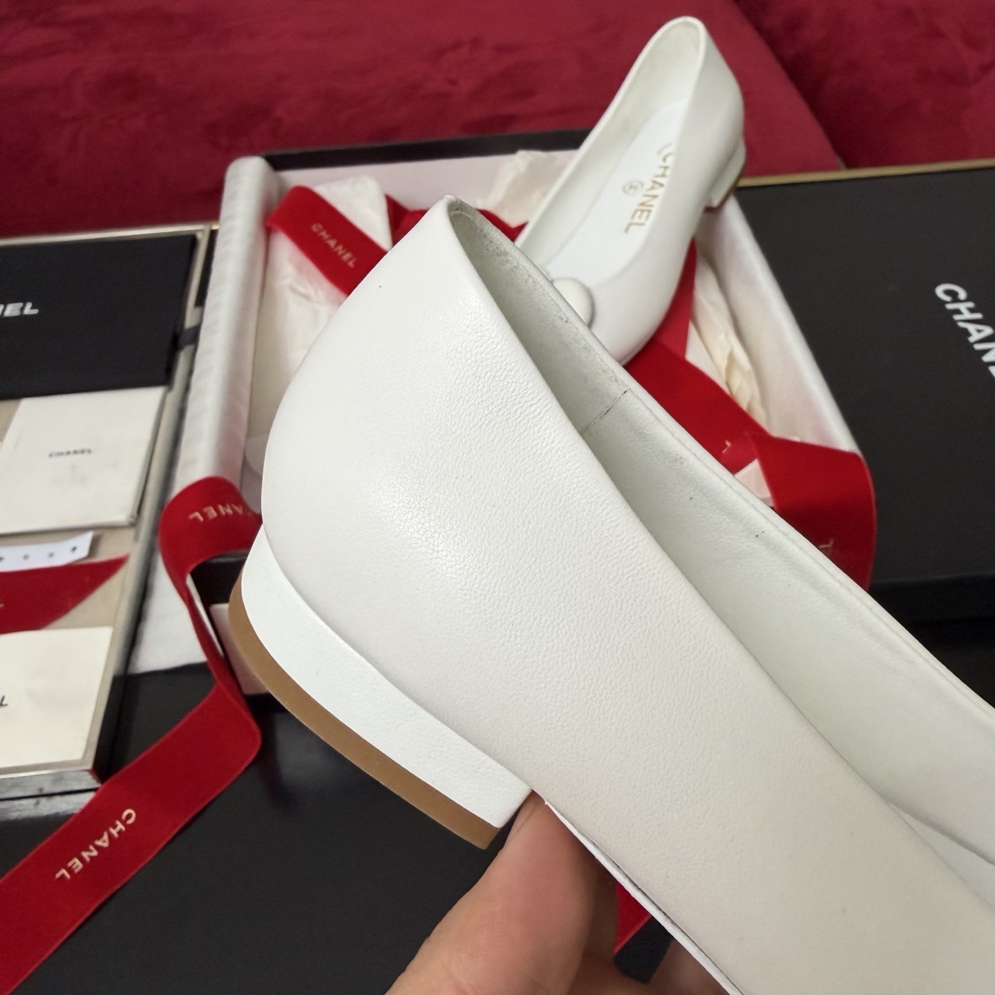 Close-up of Chanel white ballet flats featuring a leather camellia flower and gold CC logo inside a luxury gift box with red ribbon. - Hình 7