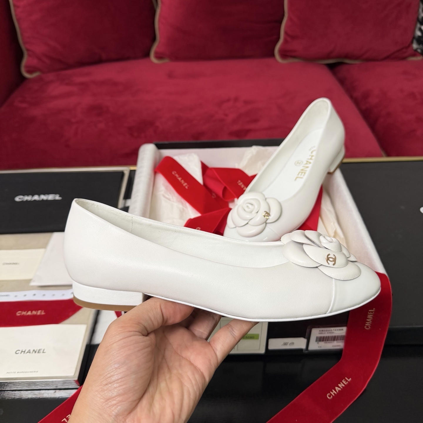 Close-up of Chanel white ballet flats featuring a leather camellia flower and gold CC logo inside a luxury gift box with red ribbon. - Hình 5
