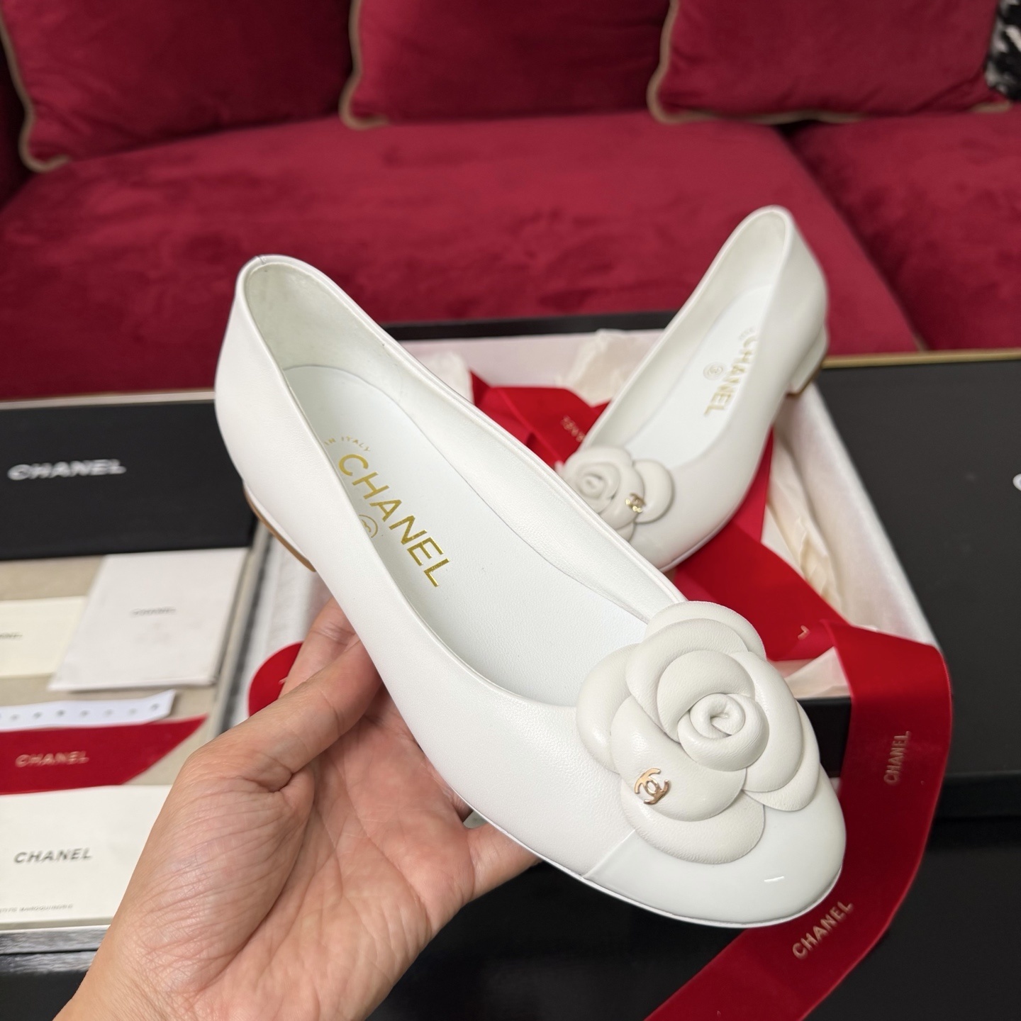 Close-up of Chanel white ballet flats featuring a leather camellia flower and gold CC logo inside a luxury gift box with red ribbon. - Hình 4