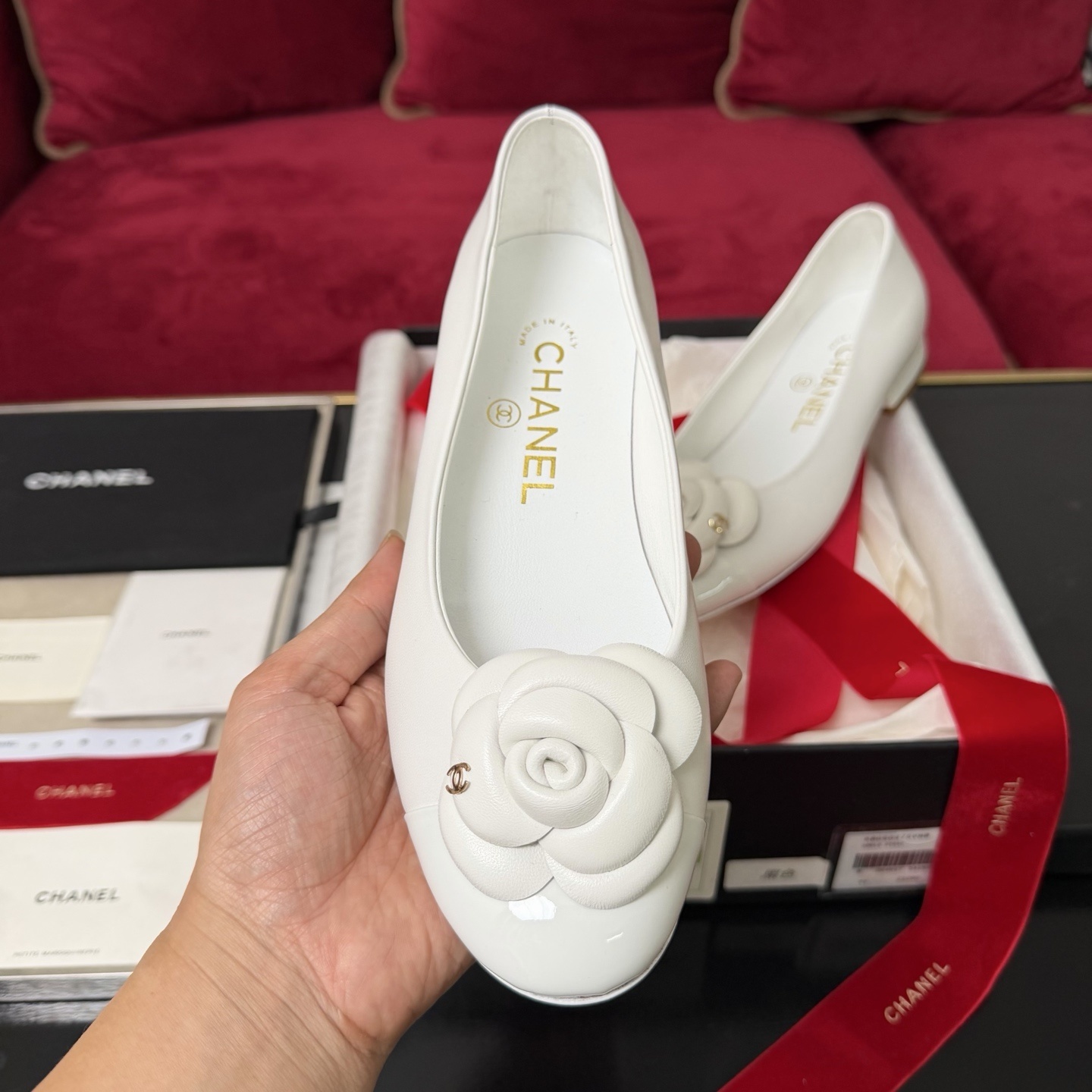 Close-up of Chanel white ballet flats featuring a leather camellia flower and gold CC logo inside a luxury gift box with red ribbon. - Hình 3
