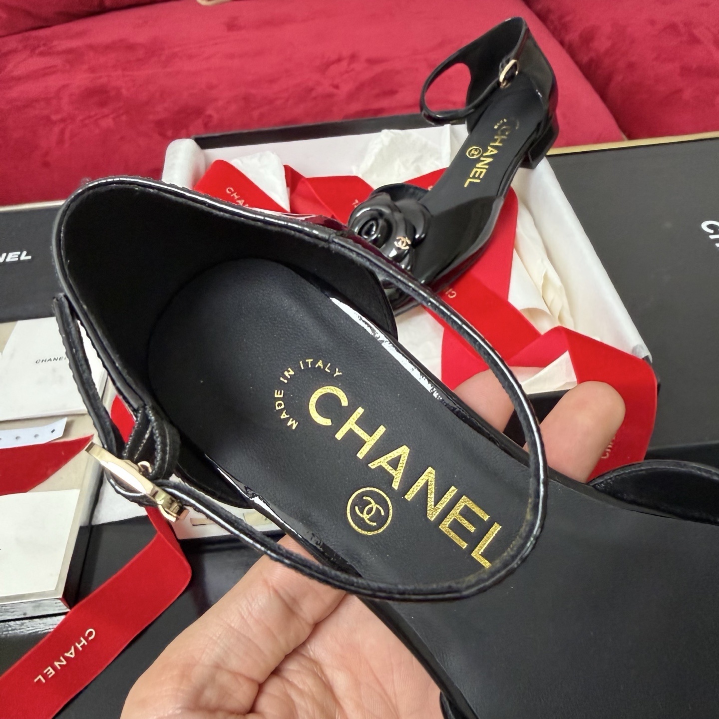A pair of black patent leather Chanel flats with a large camellia flower on the toe and an ankle strap, displayed in a Chanel box with red ribbons. - Hình 8