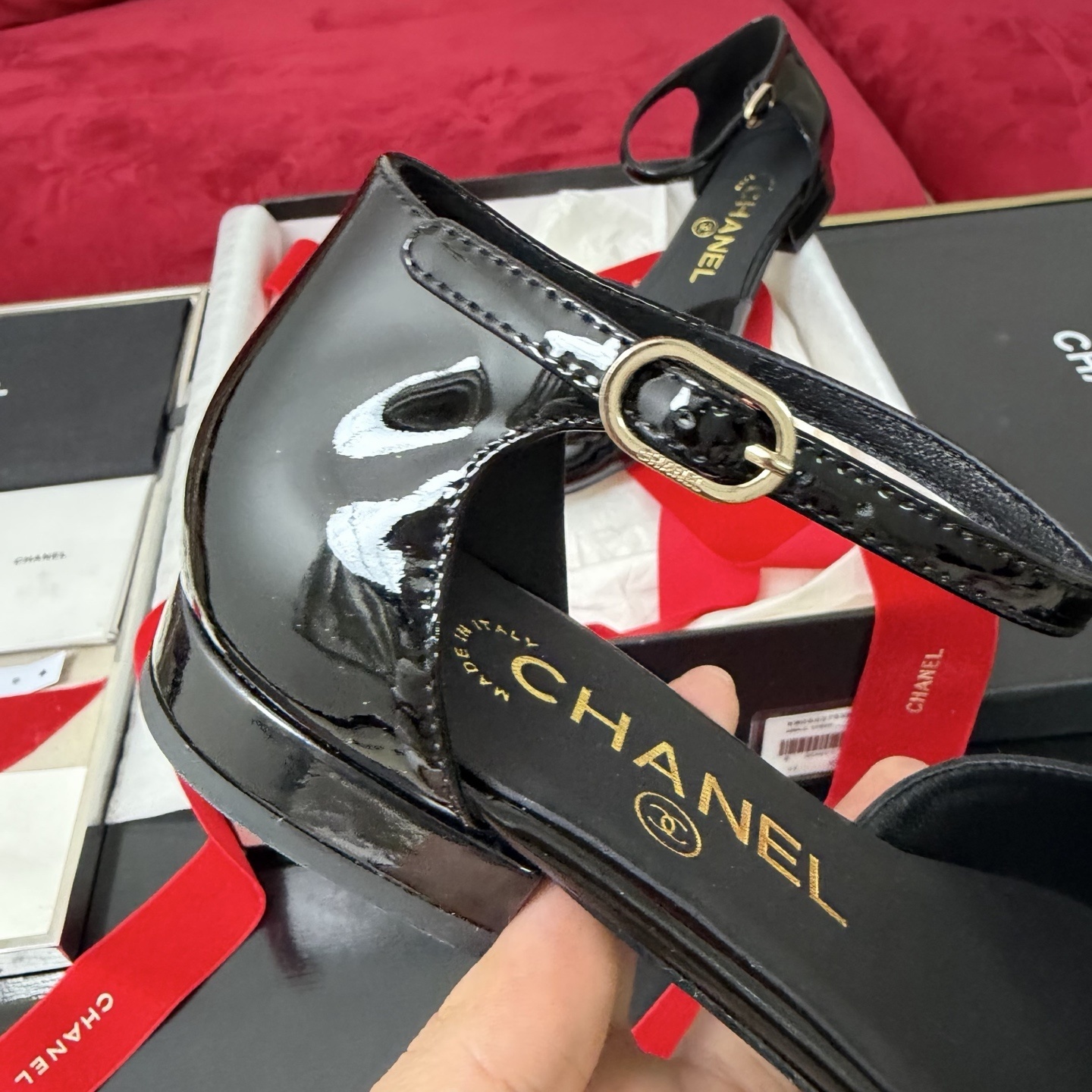 A pair of black patent leather Chanel flats with a large camellia flower on the toe and an ankle strap, displayed in a Chanel box with red ribbons. - Hình 7