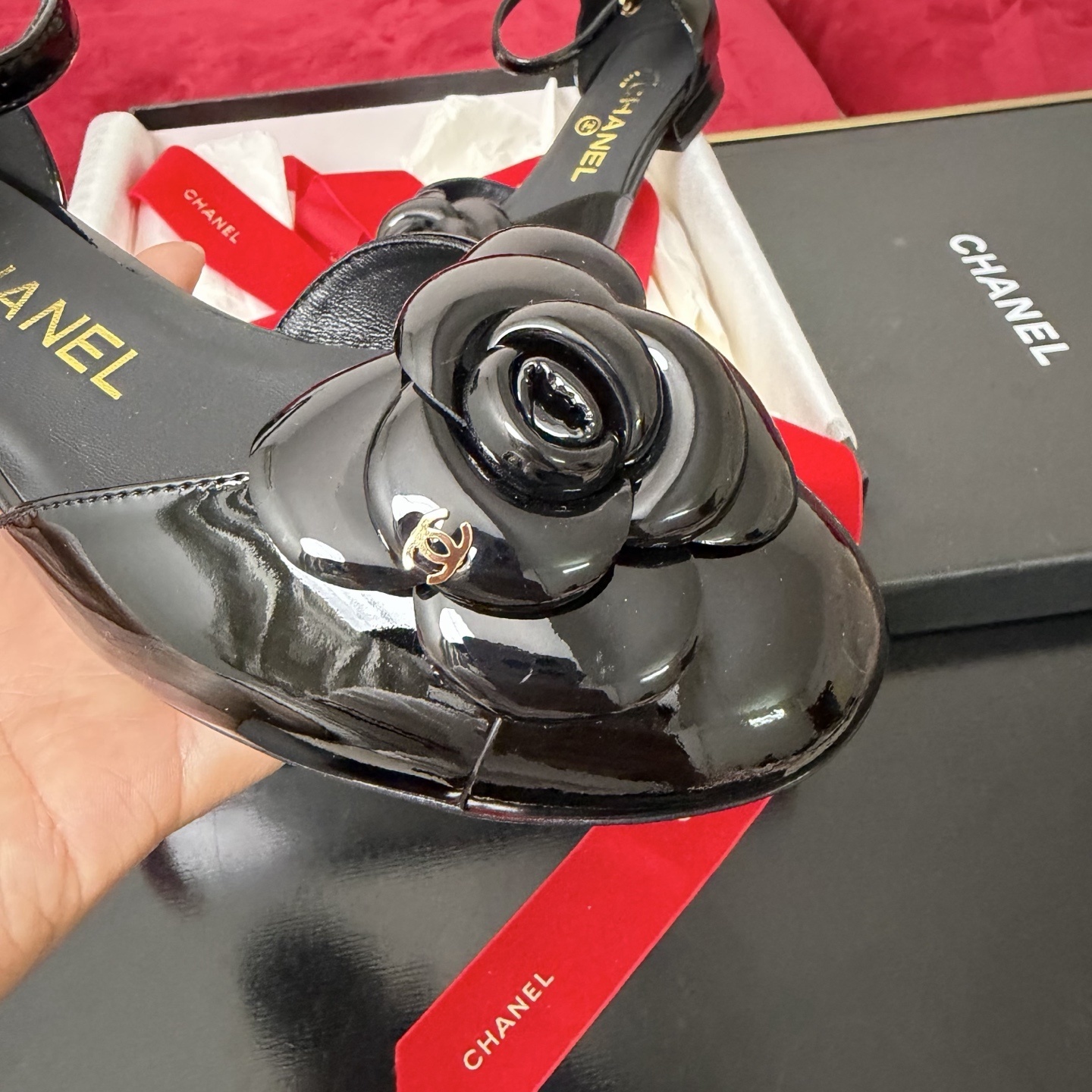 A pair of black patent leather Chanel flats with a large camellia flower on the toe and an ankle strap, displayed in a Chanel box with red ribbons. - Hình 6