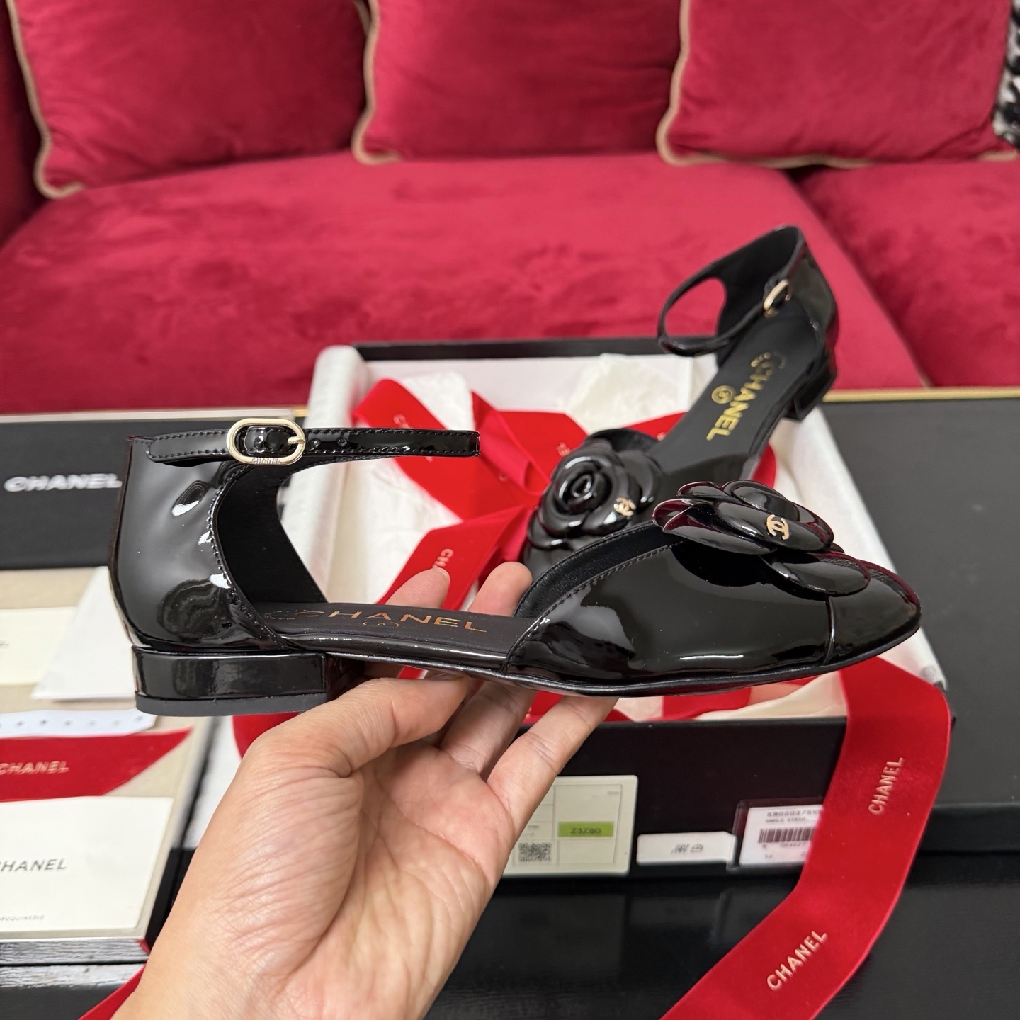 A pair of black patent leather Chanel flats with a large camellia flower on the toe and an ankle strap, displayed in a Chanel box with red ribbons. - Hình 5