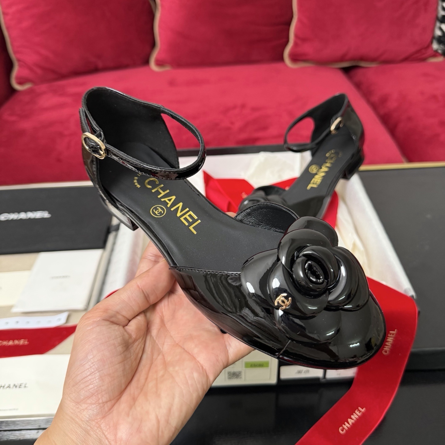 A pair of black patent leather Chanel flats with a large camellia flower on the toe and an ankle strap, displayed in a Chanel box with red ribbons. - Hình 4