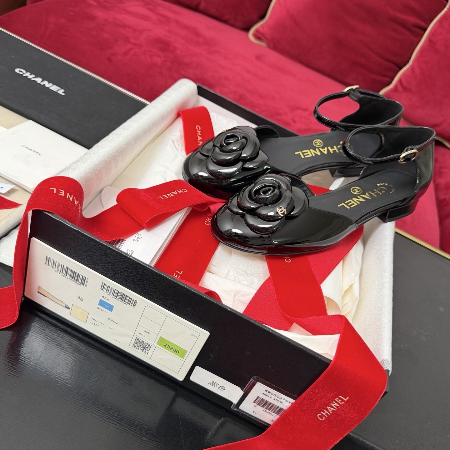 A pair of black patent leather Chanel flats with a large camellia flower on the toe and an ankle strap, displayed in a Chanel box with red ribbons. - Hình 2