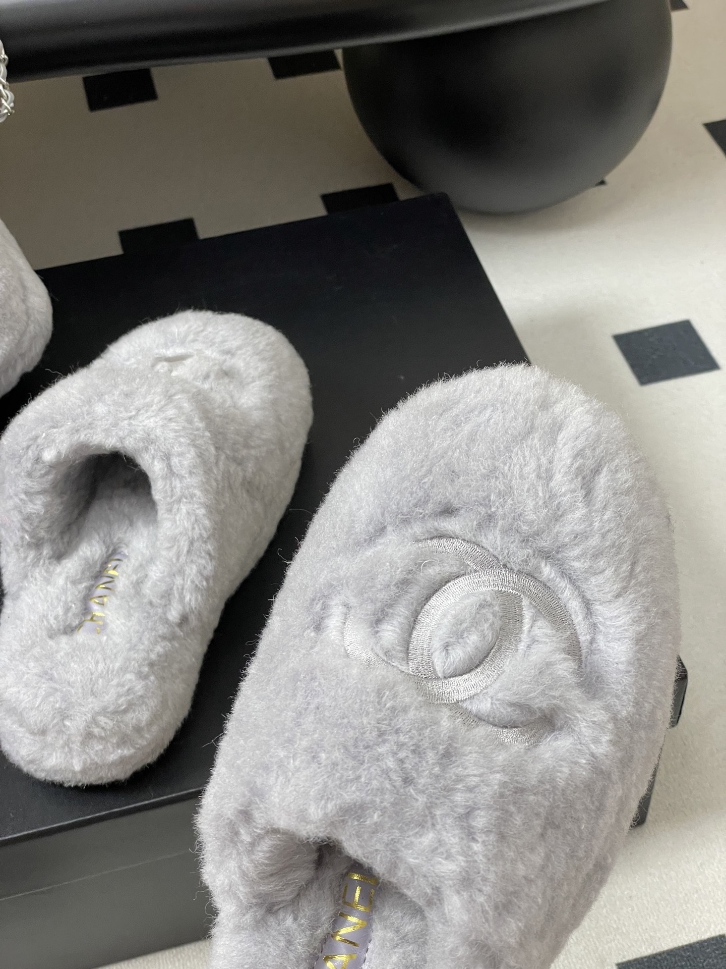 A pair of plush grey faux fur indoor slippers with a white embroidered interlocking circular logo on the top. - Hình 8