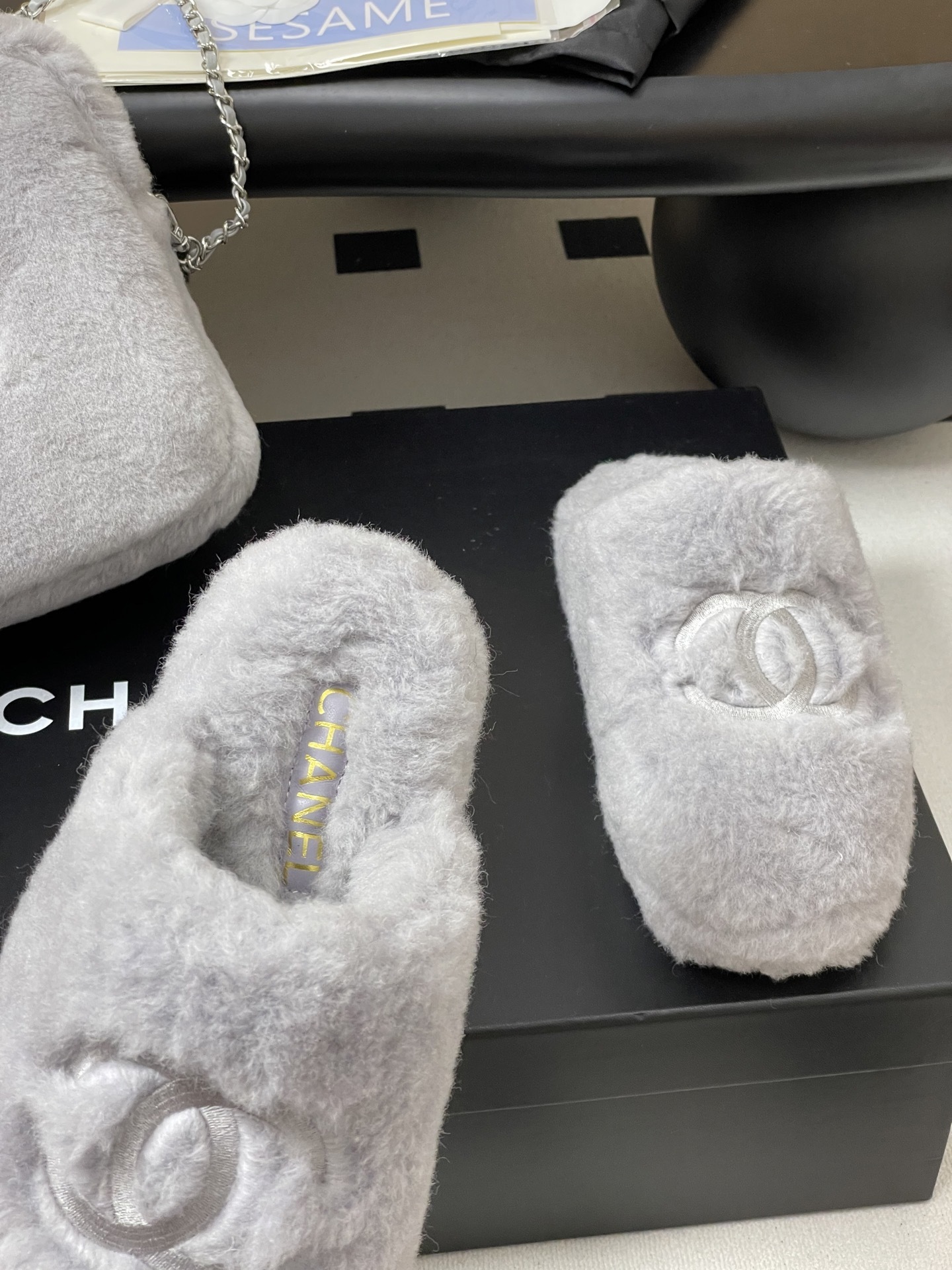 A pair of plush grey faux fur indoor slippers with a white embroidered interlocking circular logo on the top. - Hình 6