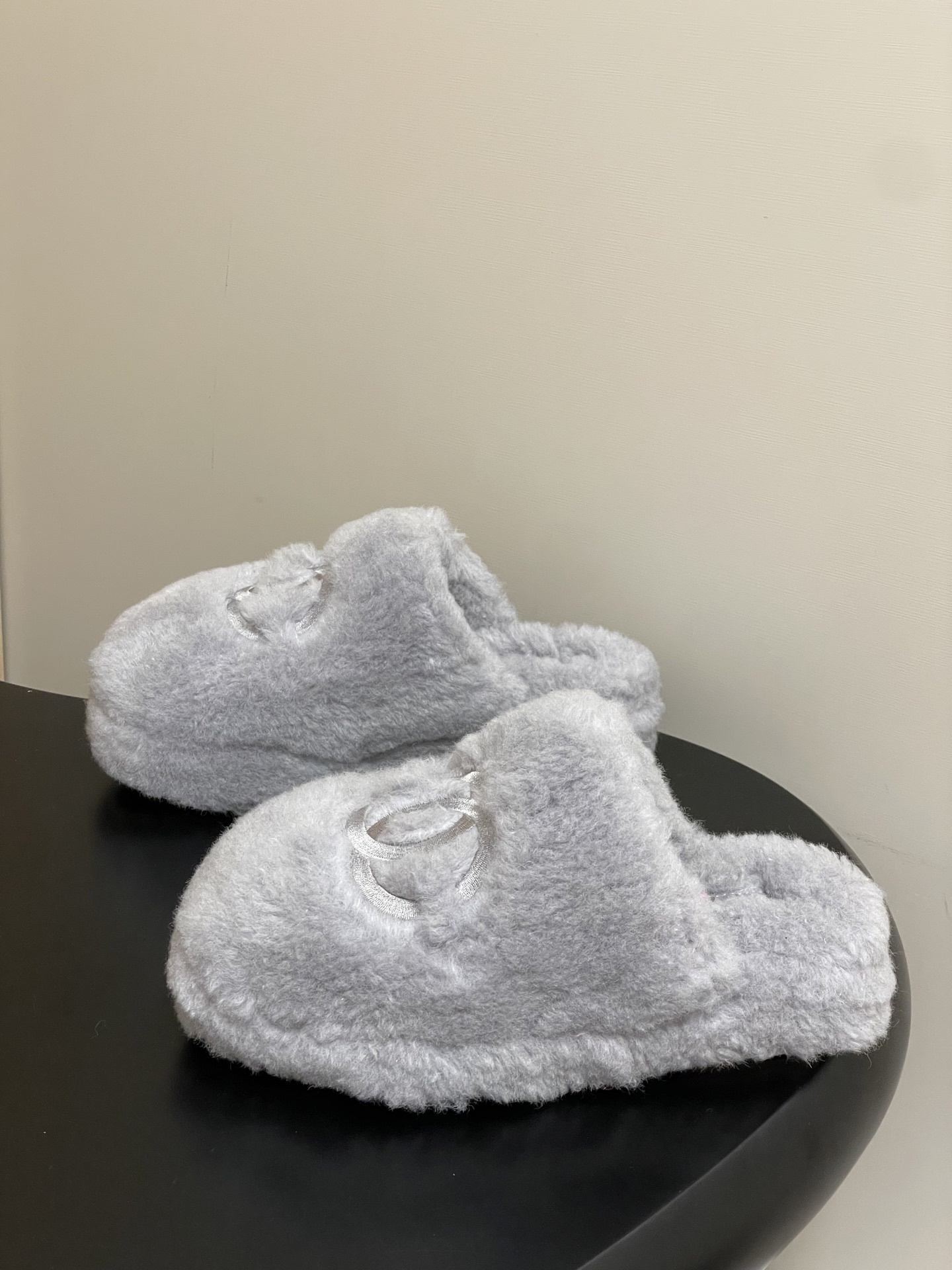 A pair of plush grey faux fur indoor slippers with a white embroidered interlocking circular logo on the top. - Hình 3