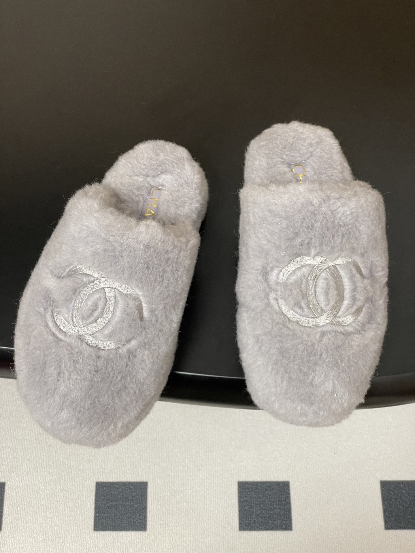 A pair of plush grey faux fur indoor slippers with a white embroidered interlocking circular logo on the top. - Hình 2