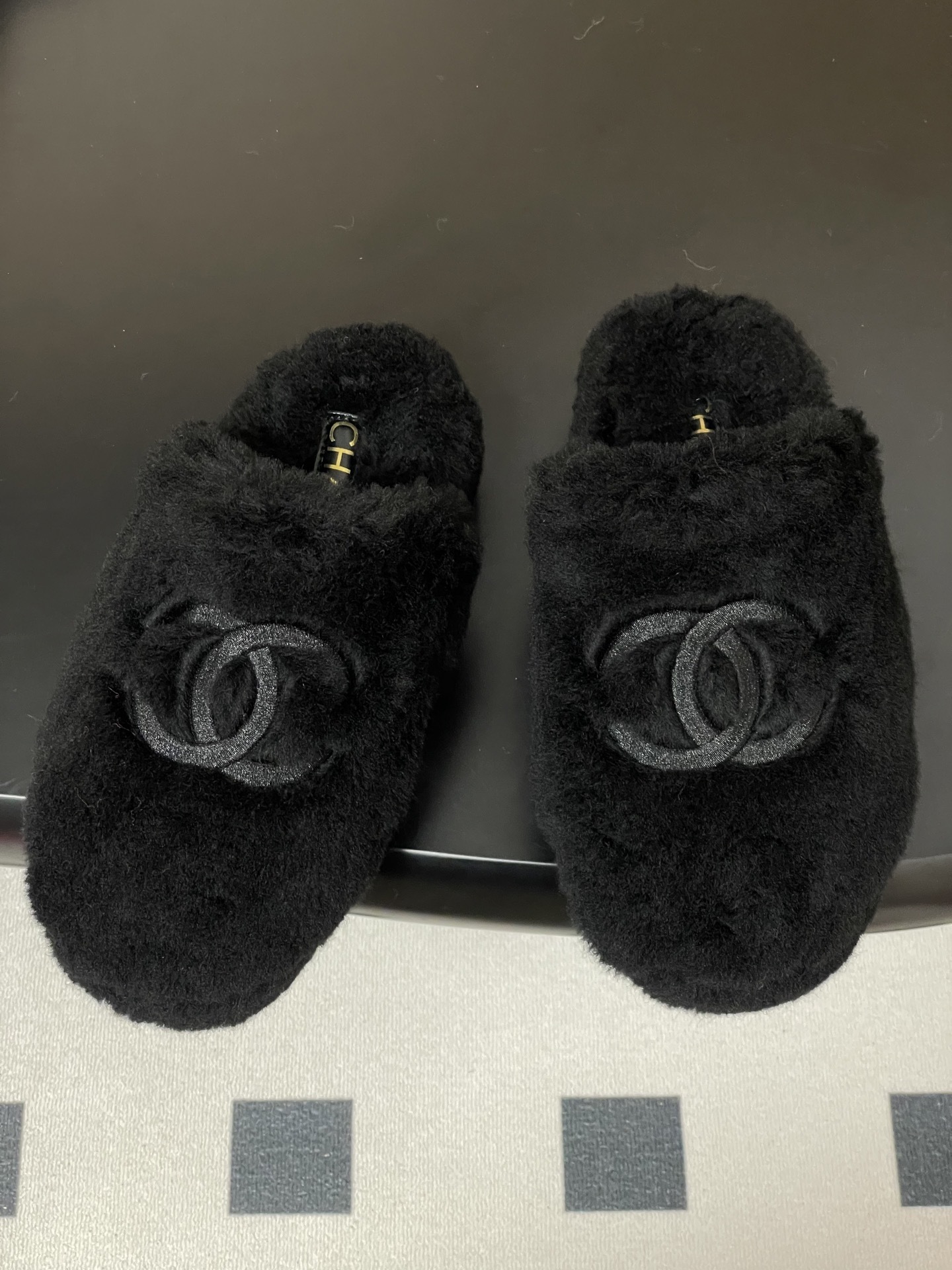 Pair of black plush fuzzy slippers with a grey interlocking CC logo on a black surface - Hình 2