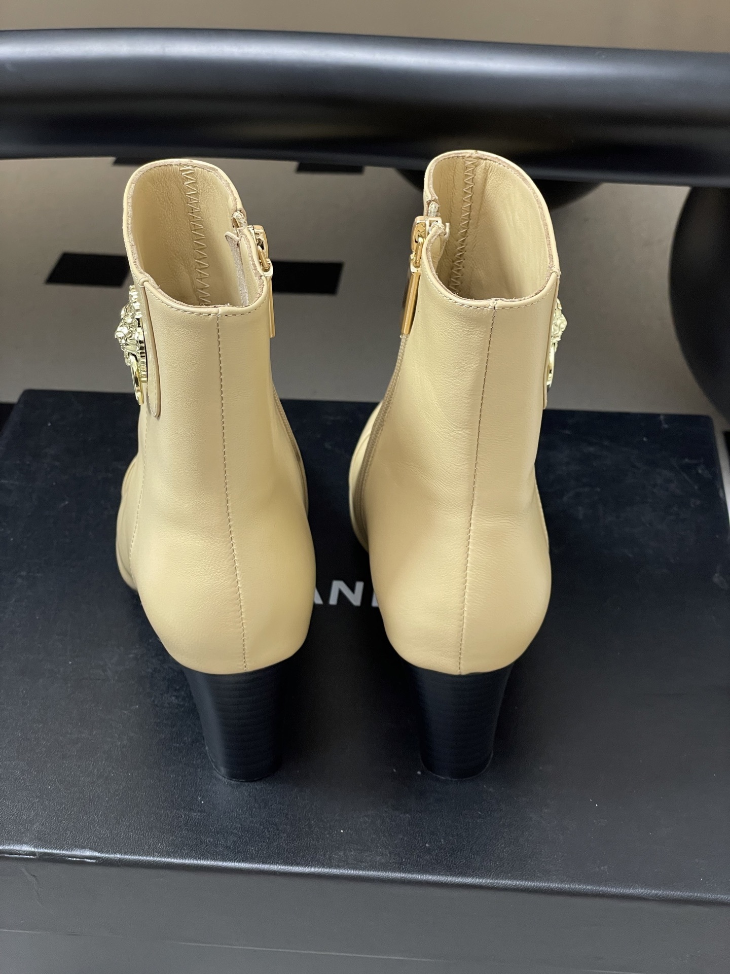 Pair of elegant beige leather ankle boots with a gold lion head embellishment and pointed toe on a black surface. - Hình 9