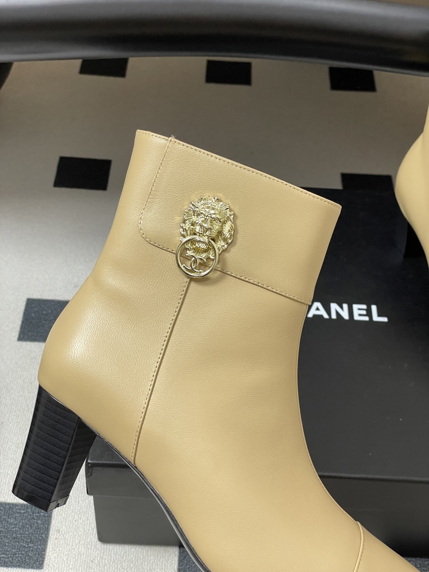 Pair of elegant beige leather ankle boots with a gold lion head embellishment and pointed toe on a black surface. - Hình 8
