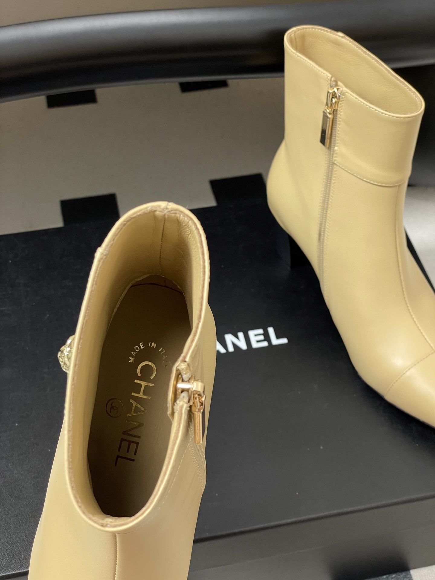 Pair of elegant beige leather ankle boots with a gold lion head embellishment and pointed toe on a black surface. - Hình 5