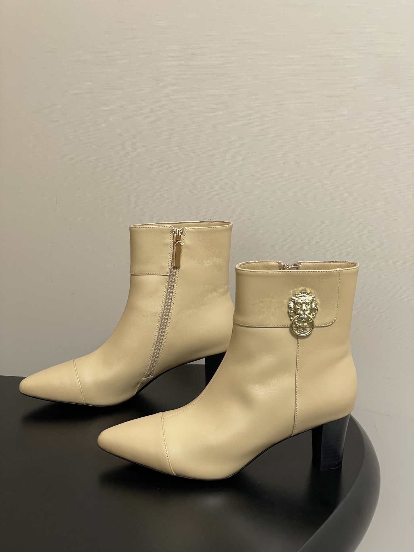 Pair of elegant beige leather ankle boots with a gold lion head embellishment and pointed toe on a black surface. - Hình 3