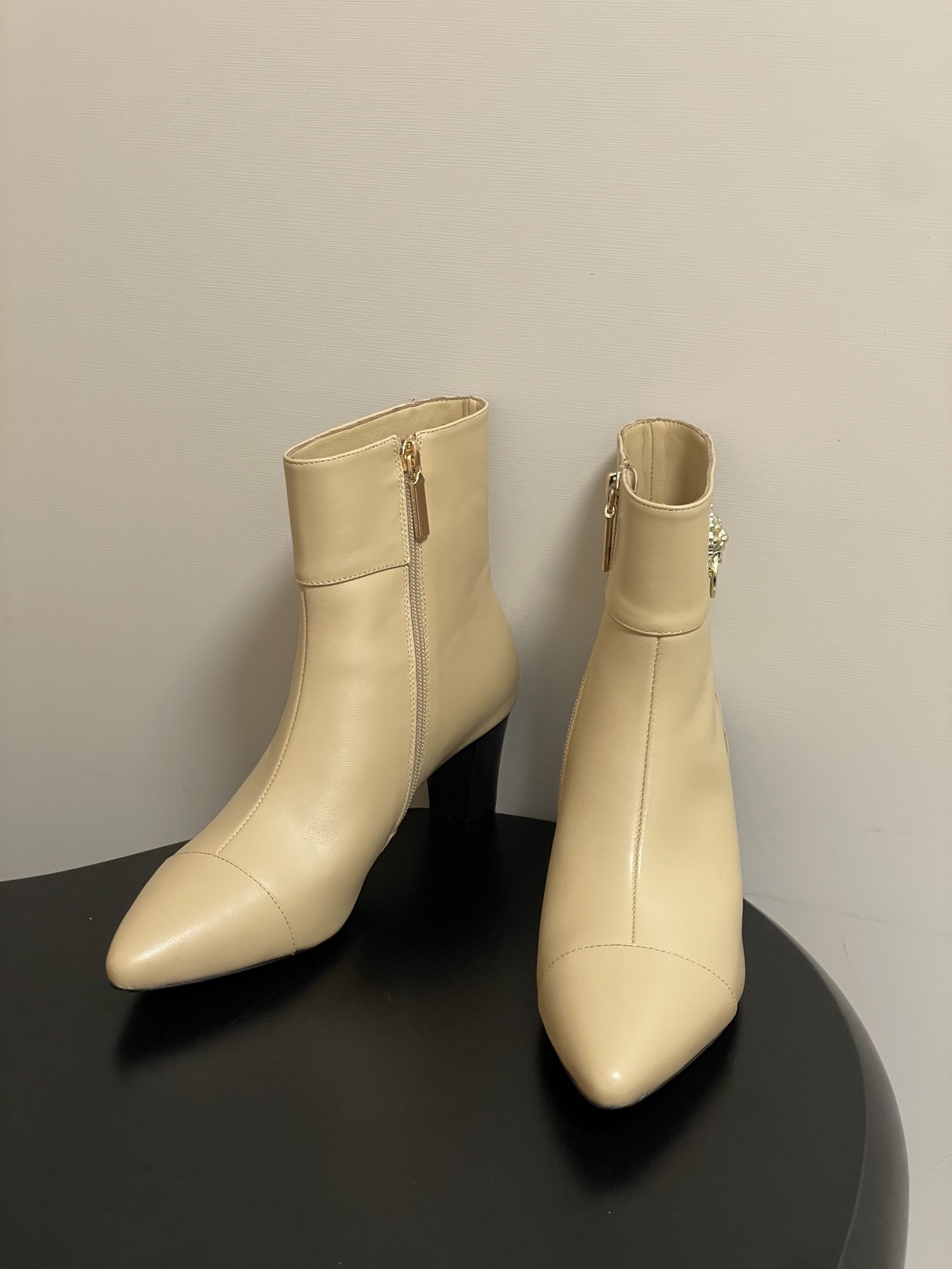 Pair of elegant beige leather ankle boots with a gold lion head embellishment and pointed toe on a black surface. - Hình 2