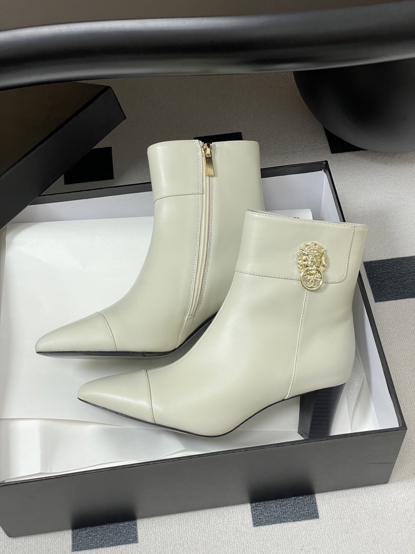 Pair of creamy white leather ankle boots with gold lion head hardware and pointed toe on a black surface - Hình 4
