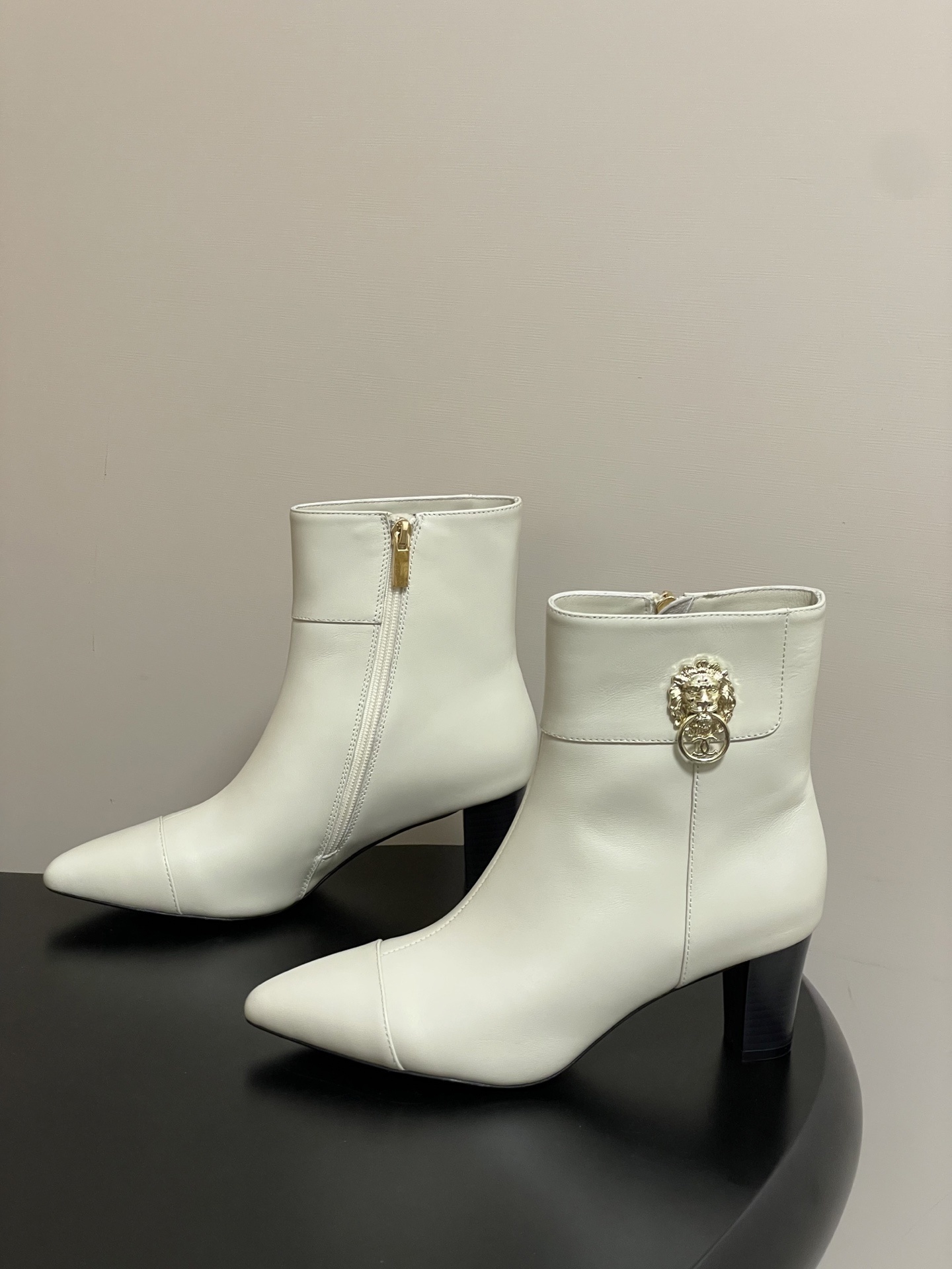 Pair of creamy white leather ankle boots with gold lion head hardware and pointed toe on a black surface - Hình 3