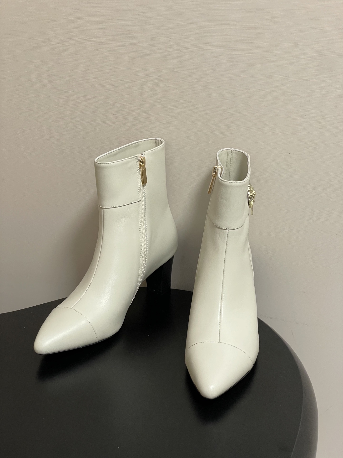 Pair of creamy white leather ankle boots with gold lion head hardware and pointed toe on a black surface - Hình 2