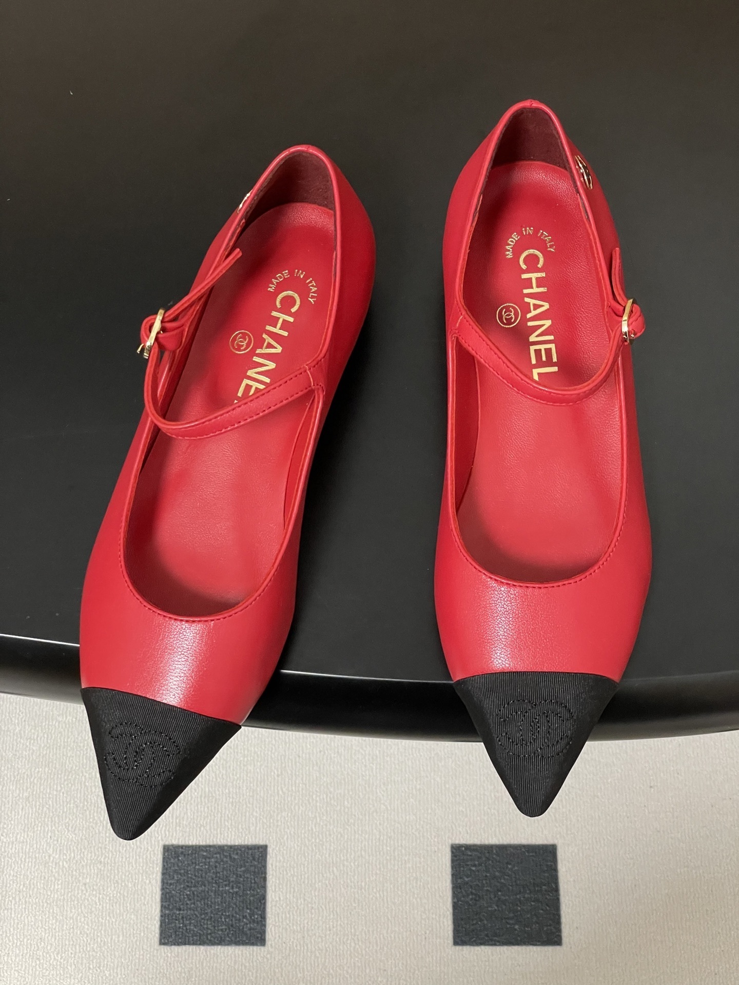 Pair of red Chanel Mary Jane leather flats with black pointed toe caps and gold CC logo on a black surface. - Hình 2