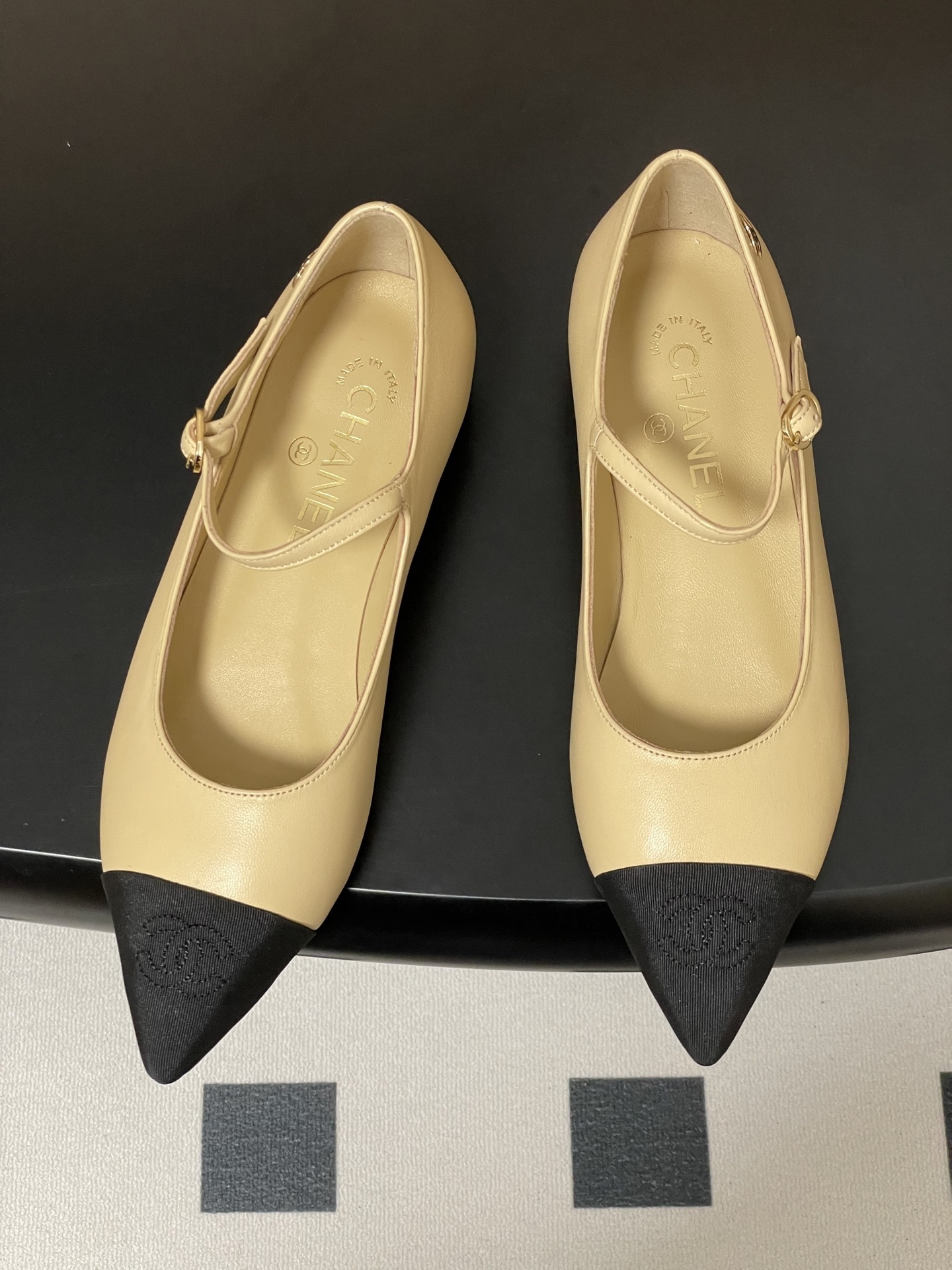 Chanel beige leather Mary Jane flats with black pointed cap toes and gold CC logo hardware on a black table. - Hình 2
