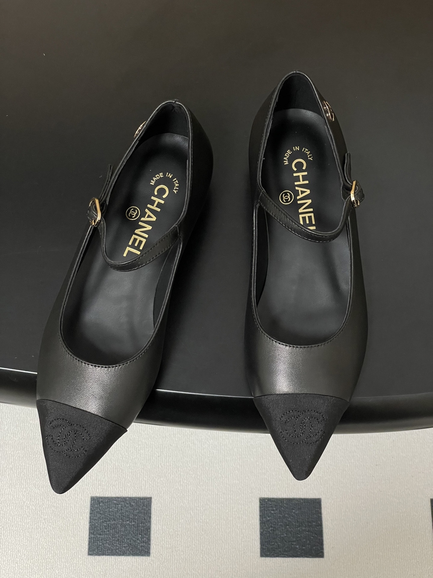 Pair of Chanel black leather Mary Jane pointed toe flats with gold CC logo and buckled strap on a black surface - Hình 2