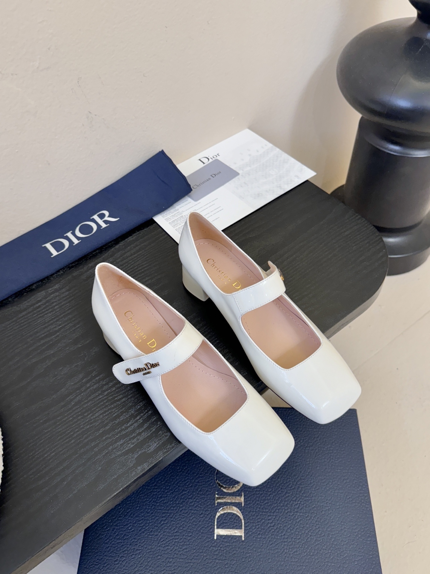 Christian Dior white patent leather Mary Jane pumps with square toe, block heel, and signature gold-tone logo strap on a black display.