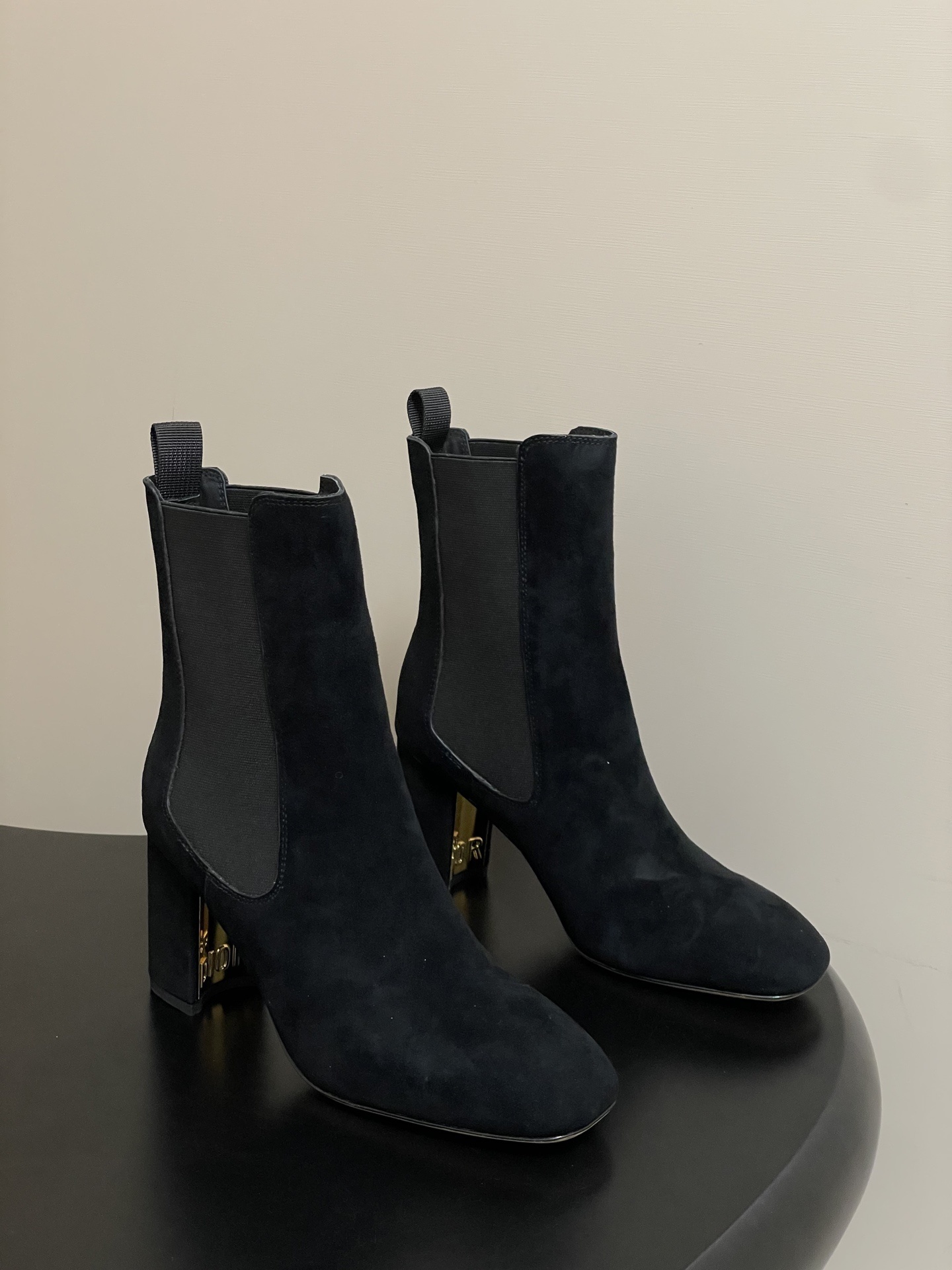 A pair of Dior black suede heeled ankle boots with gold-tone Dior logo signature on the heels, displayed on a black surface.