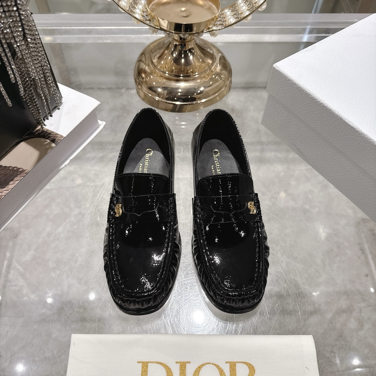 A pair of Christian Dior Boy loafers in shiny black patent calfskin featuring a gold CD logo on the strap, displayed on a glass table.